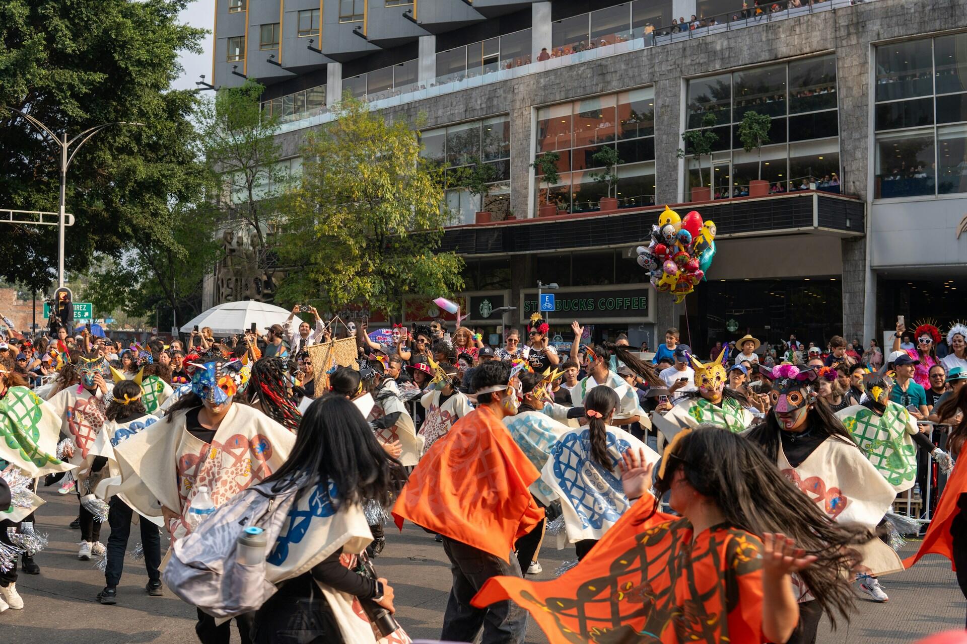 A vibrant parade scene filled with dancers in colorful costumes, masks, and banners, celebrating a festive event in an urban setting.