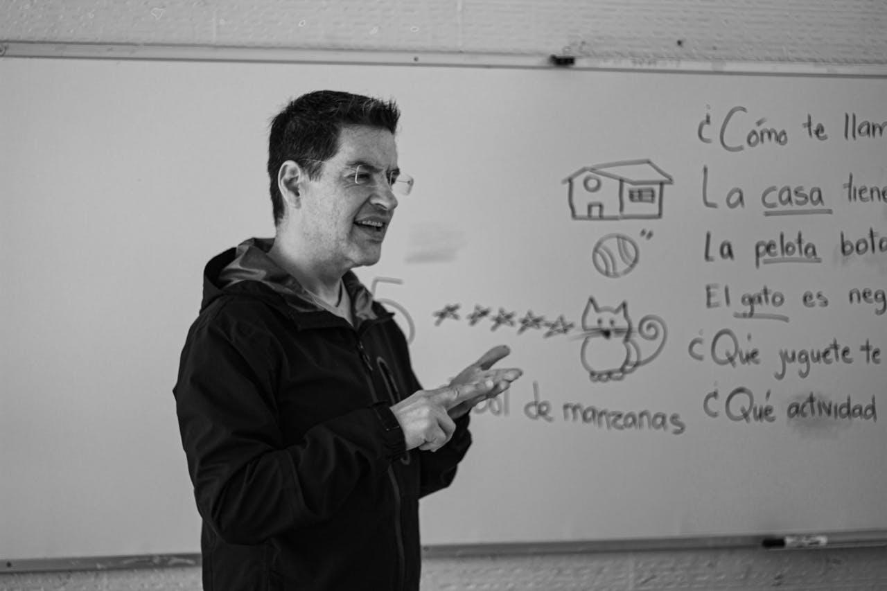 A person gestures while explaining concepts related to a cat drawing and vocabulary words on a whiteboard in a classroom setting.