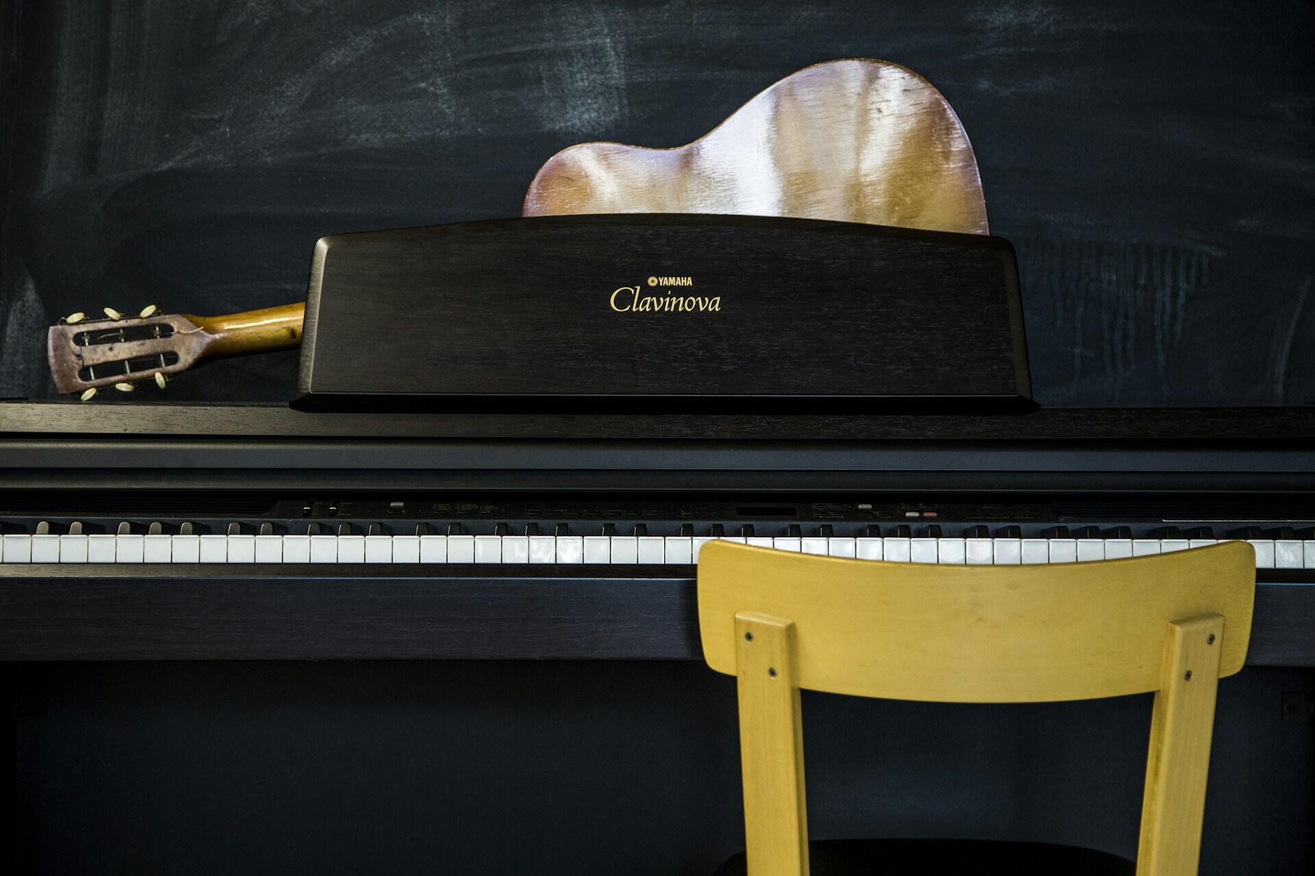 A Yamaha Clavinova digital piano sits in front of a yellow chair, with an acoustic guitar leaning against a chalkboard backdrop.