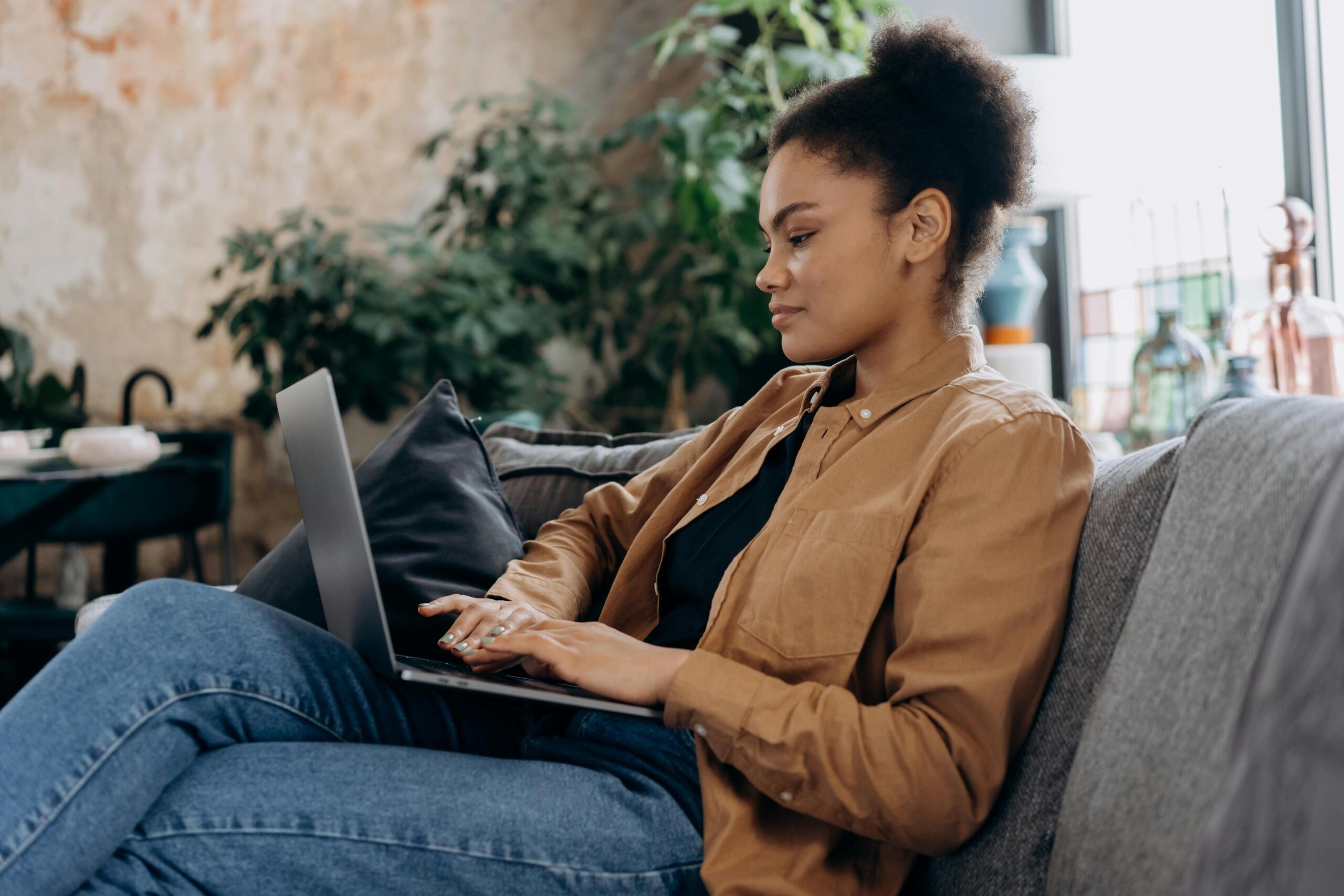 A woman in casual professional attire sitting comfortably on a couch, working on a laptop placed on her lap, appearing focused and engaged.