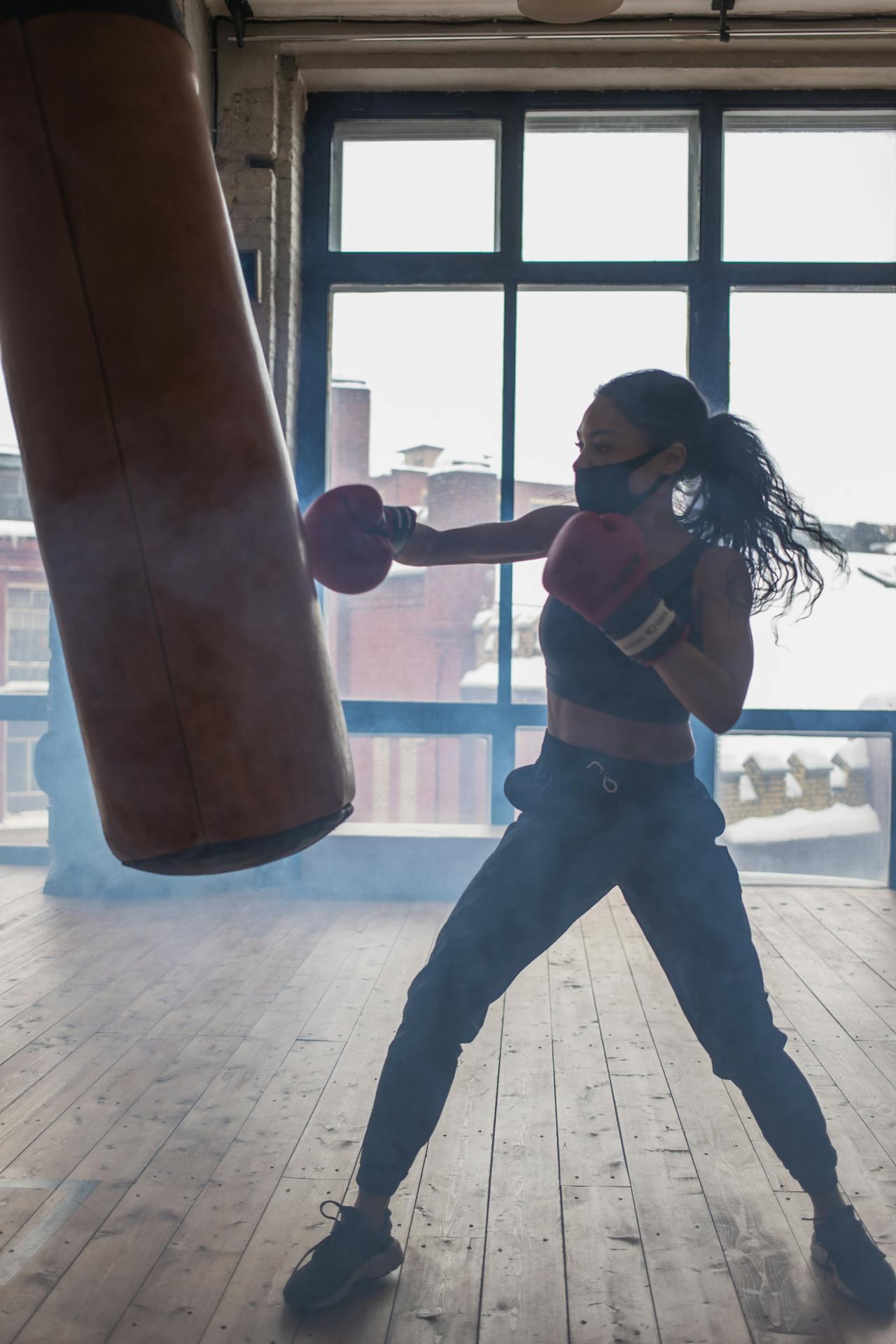 A boxer throws a punch at a heavy bag in a well-lit gym, with smoke and large windows in the background.