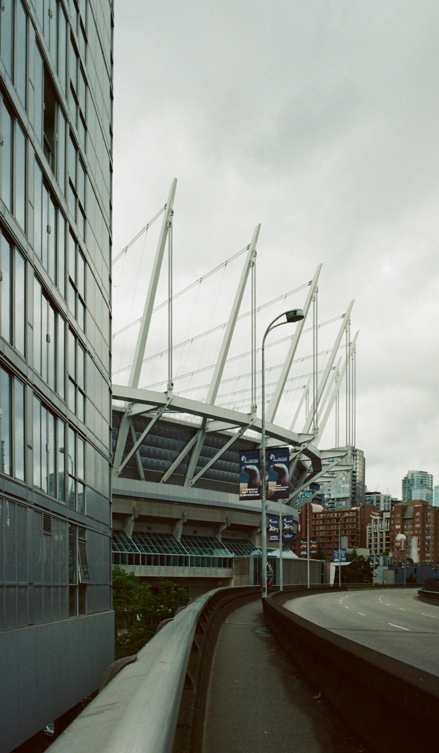 A view of a modern stadium with distinctive white supports, flanked by a reflective building and a road under a cloudy sky.