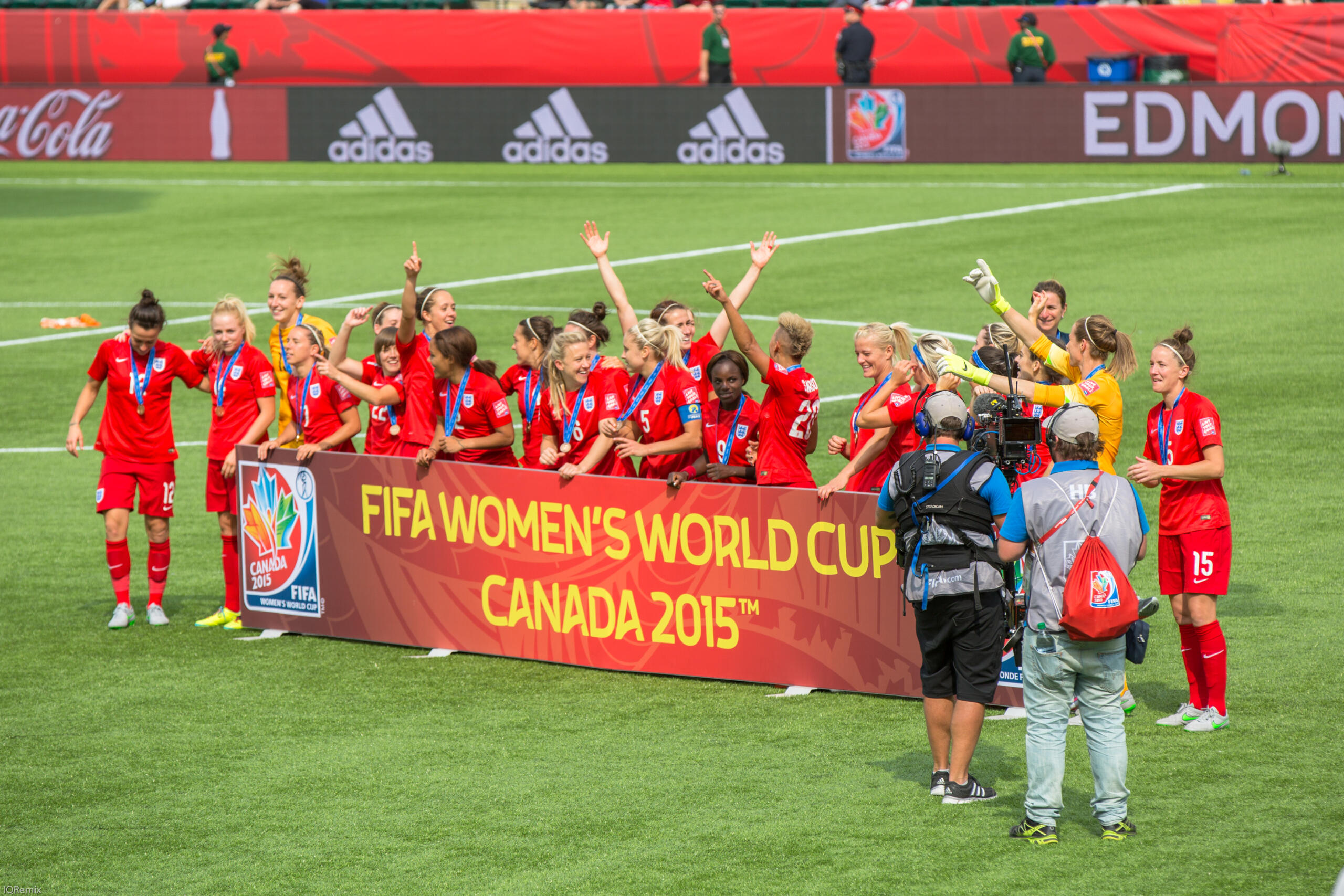 England women’s soccer team celebrating with medals behind a FIFA Women’s World Cup Canada 2015 banner on the field, with cameras filming the moment.