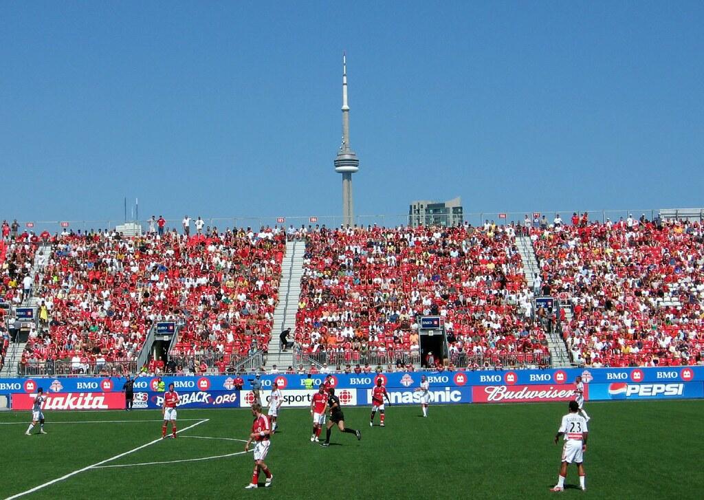 Packed stadium of Canadian soccer fans in red, with the CN Tower visible in the background.