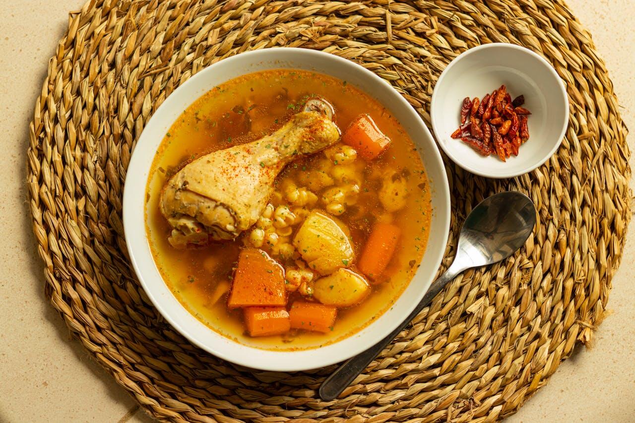 Bowl of hearty chicken soup with vegetables and corn, served with a side of dried chili peppers on a woven placemat.