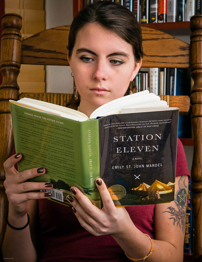A person sitting in a wooden chair reads the novel "Station Eleven" by Emily St. John Mandel, showcasing a cozy reading environment.
