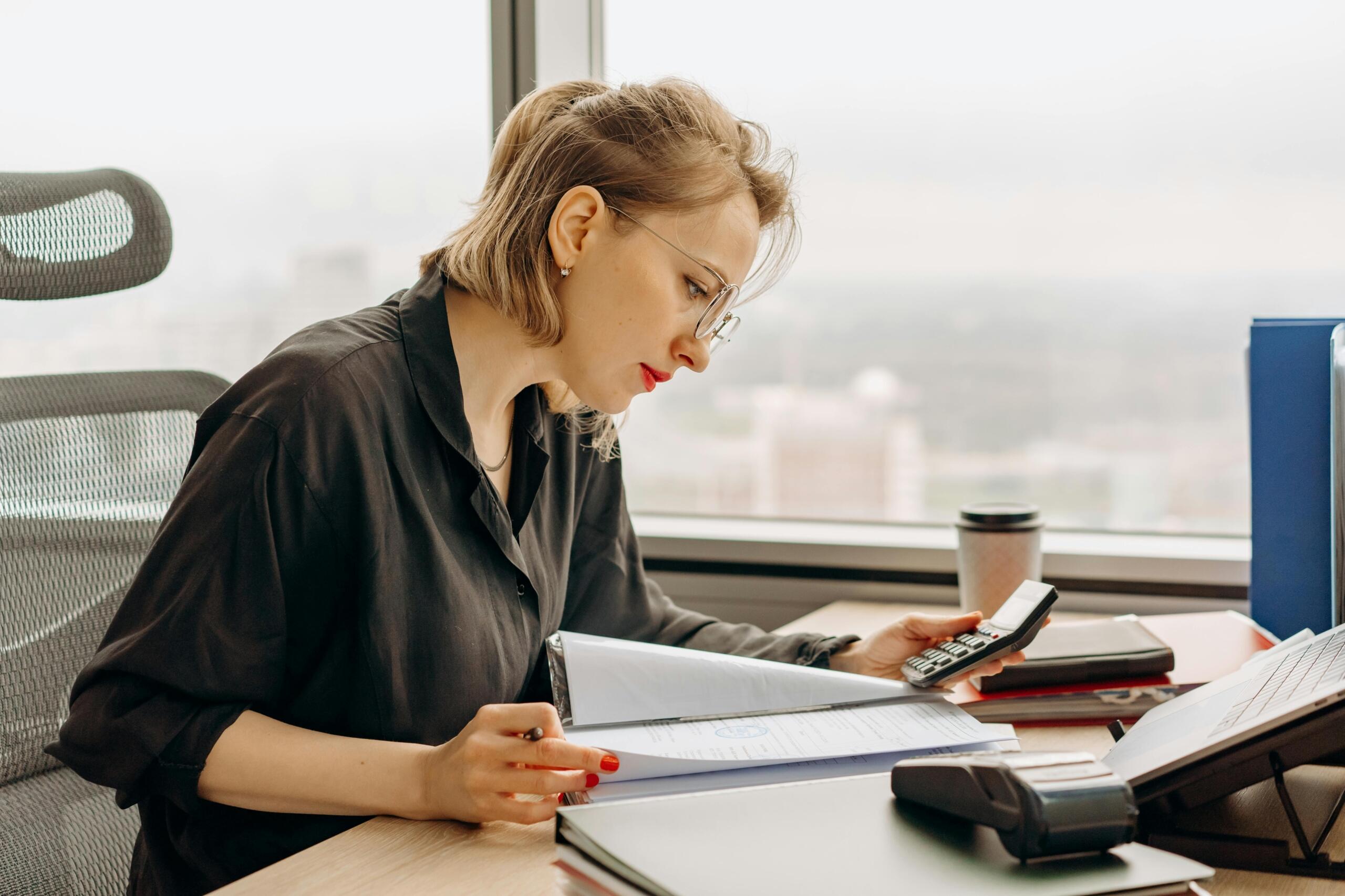 A woman sitting at a desk looking at papers while holding a calculator, with documents and a laptop in front of her in a well-lit office space.