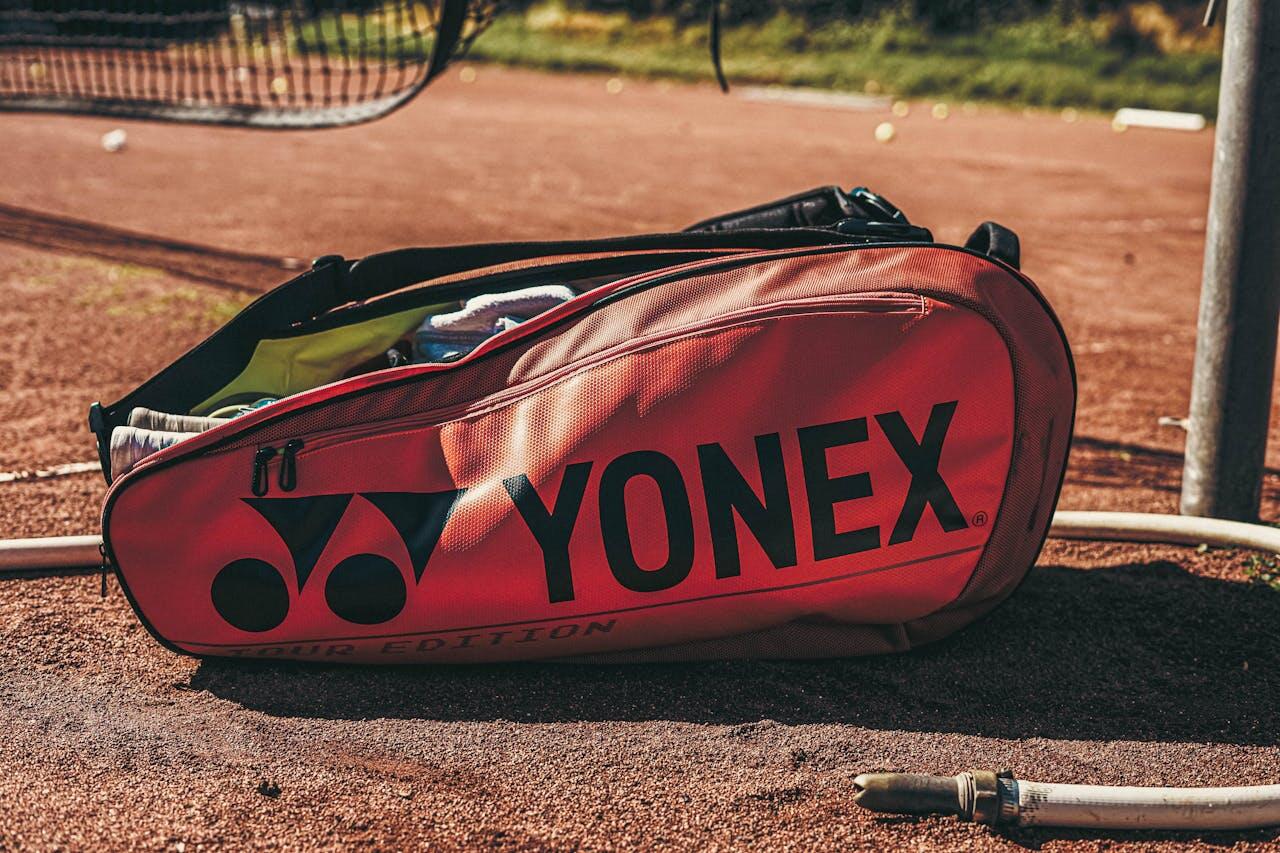 A vibrant red Yonex tennis bag rests on clay court, showcasing its logo and partially unzipped to reveal gear inside.