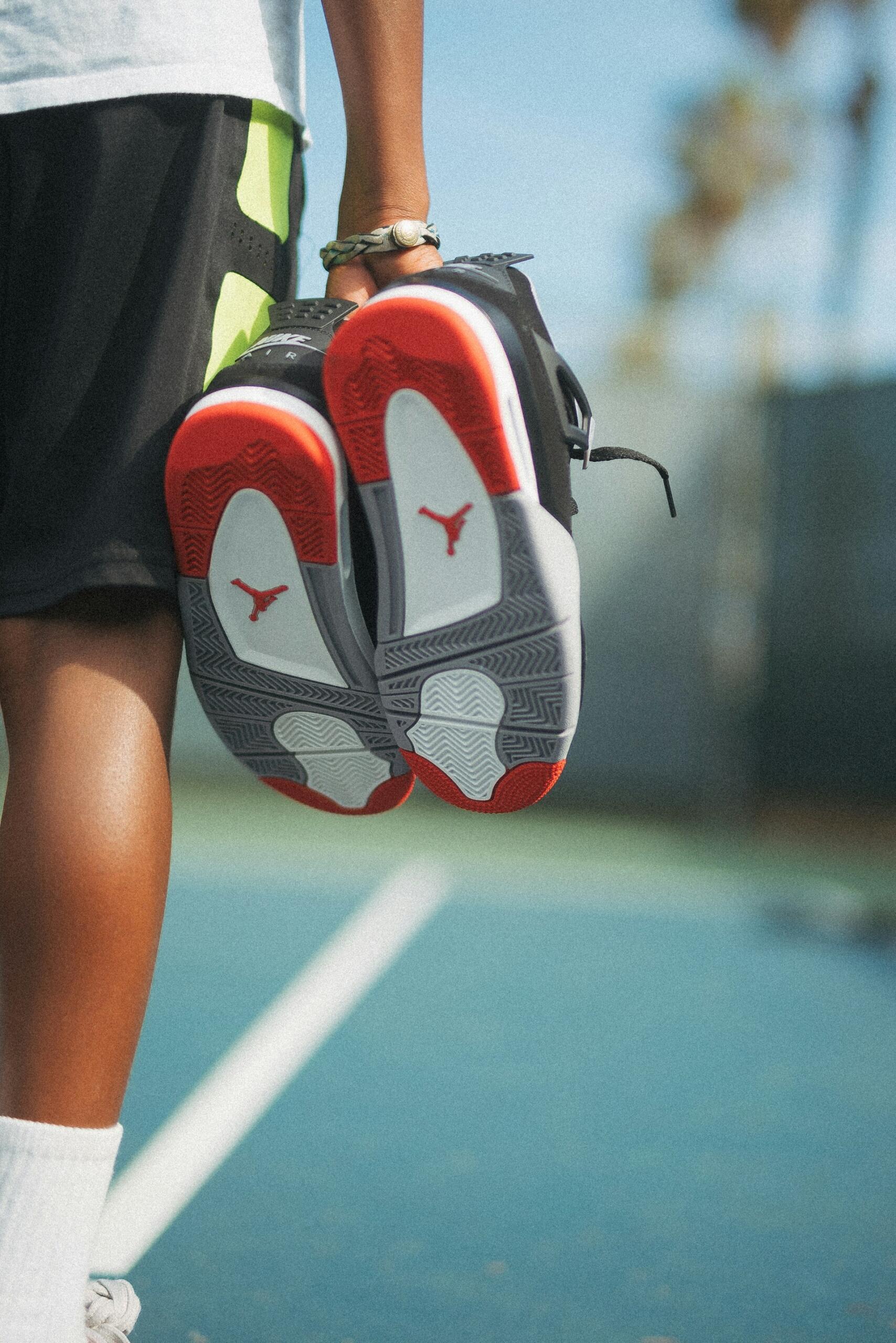 A close-up of a person holding black and red Jordan sneakers, with the tennis court and palm trees blurred in the background.