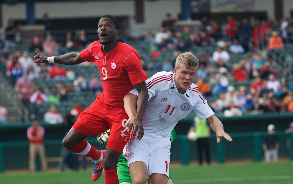 Canadian player Tosaint Ricketts and Denmark’s Andreas Cornelius compete for the ball during an international soccer match, with a full crowd in the background.