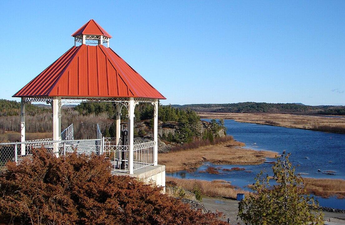 A gazebo with a red roof overlooks a calm river surrounded by golden marshlands and distant green hills under a clear blue sky.