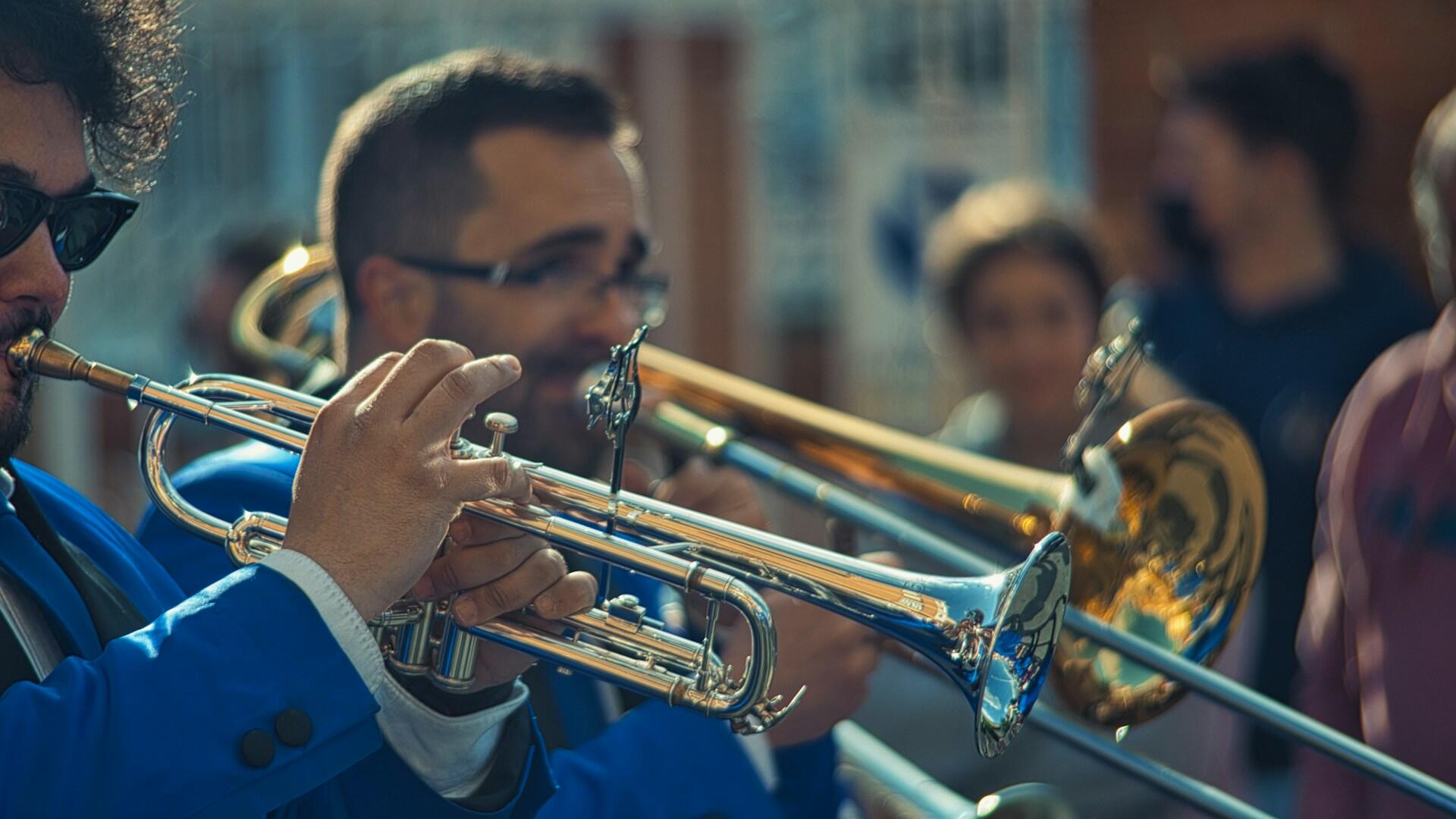 A group of musicians in blue jackets play brass instruments, including trumpets and trombones, in a lively outdoor setting.
