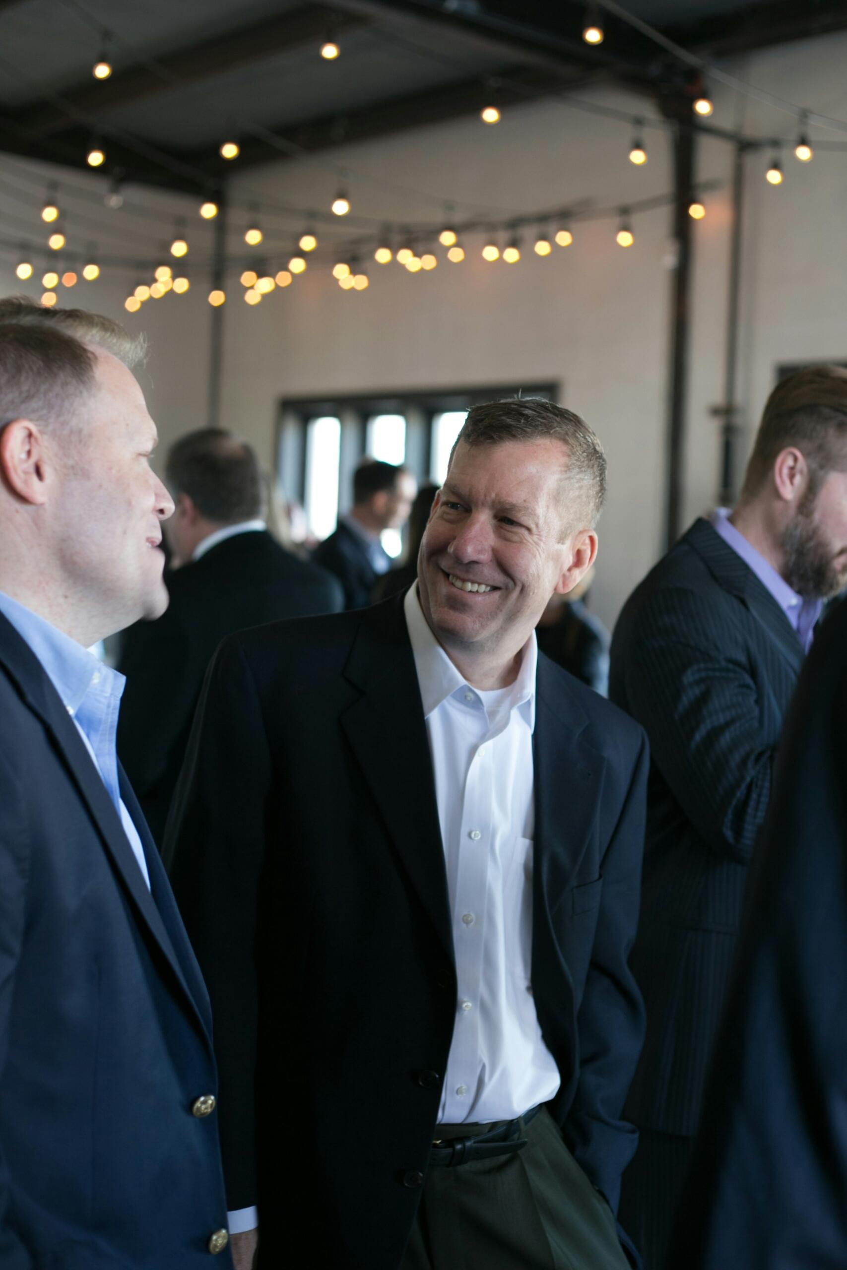 Two men in business attire share a conversation and a laugh at a professional networking event, with string lights and other attendees in the background.