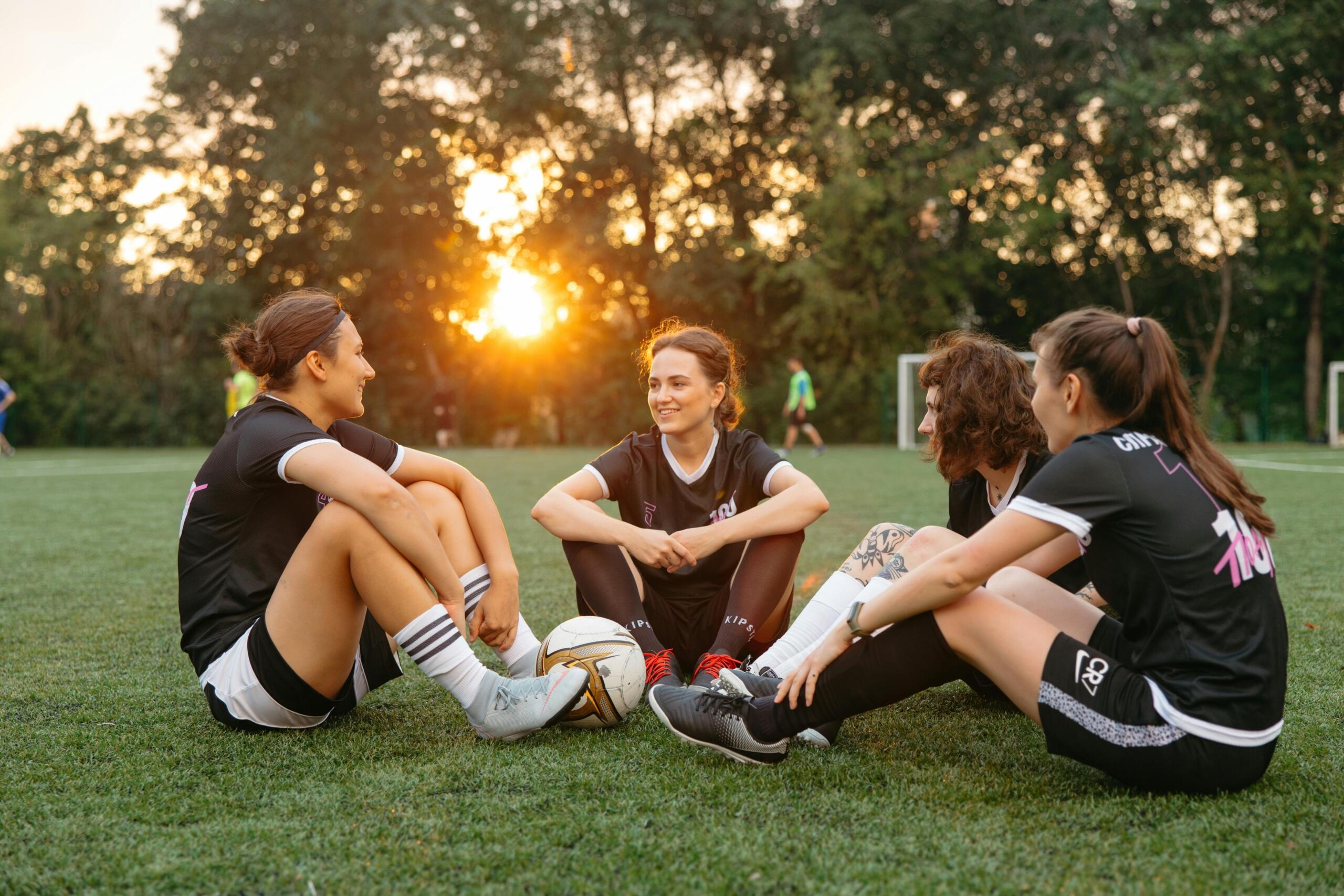 Three women in soccer uniforms sitting on a soccer field, engaged in conversation.