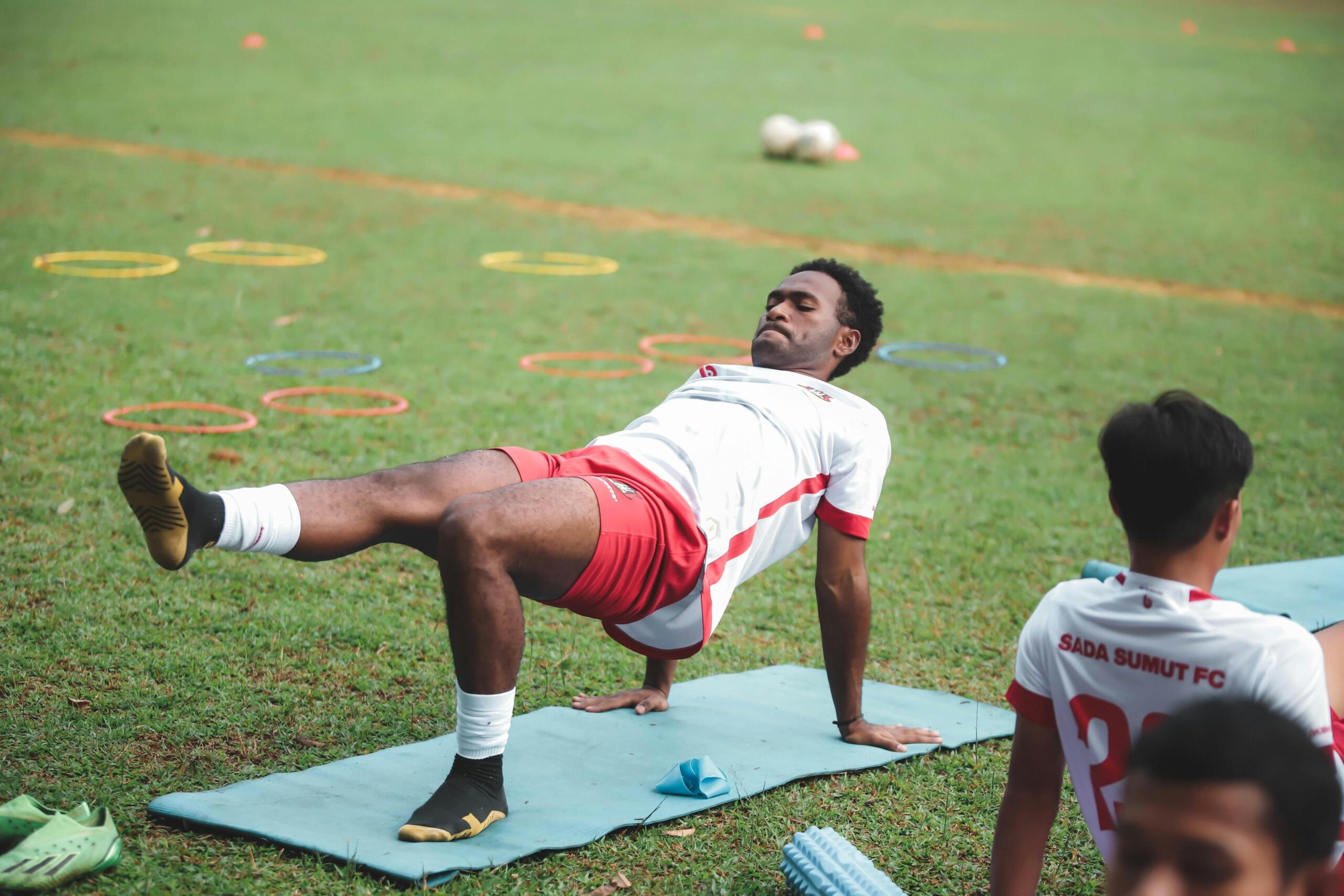 A soccer player stretching on a mat placed on a grass field.