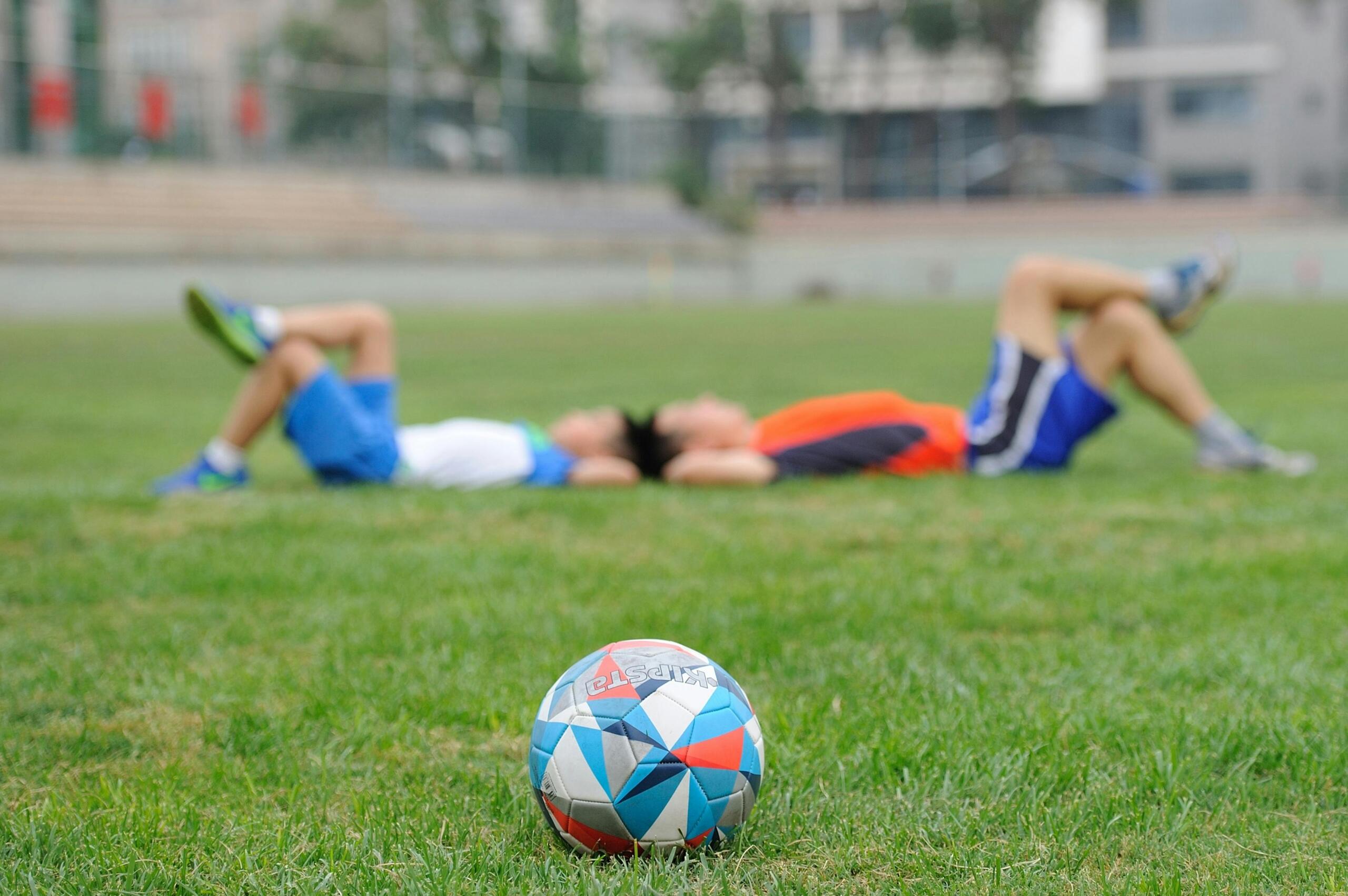 Two soccer players lying on a grass field with a soccer ball in the foreground. 