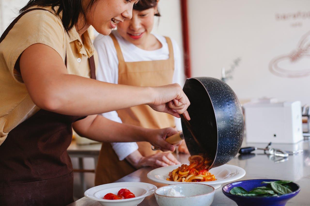 Two people in aprons are preparing a dish in a kitchen, pouring pasta with sauce onto a plate, surrounded by ingredients.