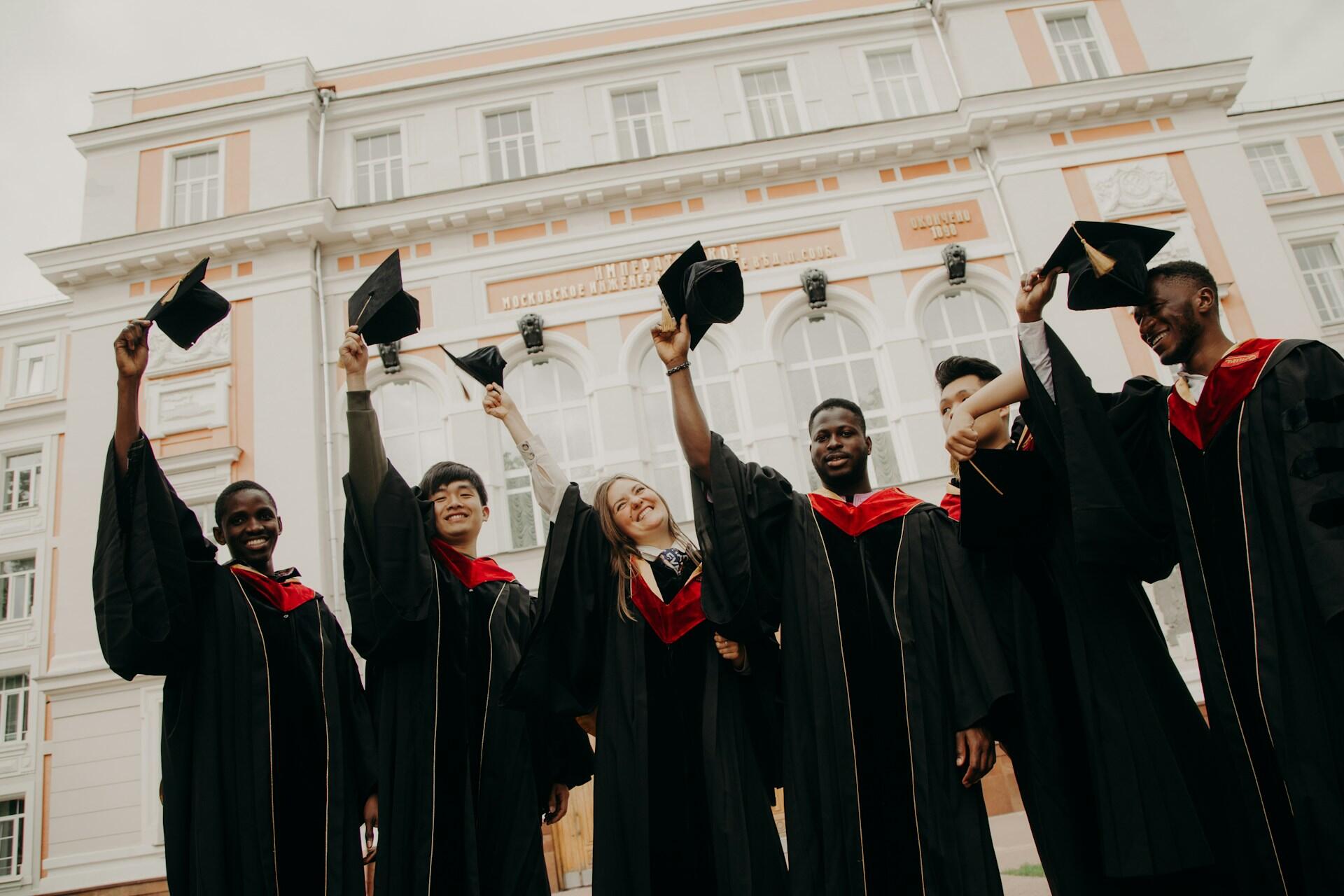 A group of graduates in black gowns and red hoods celebrate by raising their caps in front of an academic building.