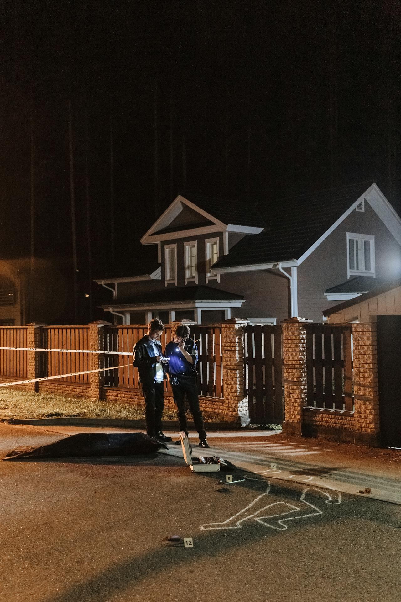 Two officers investigate a crime scene at night, marked by evidence markers and a chalk outline in front of a suburban home.