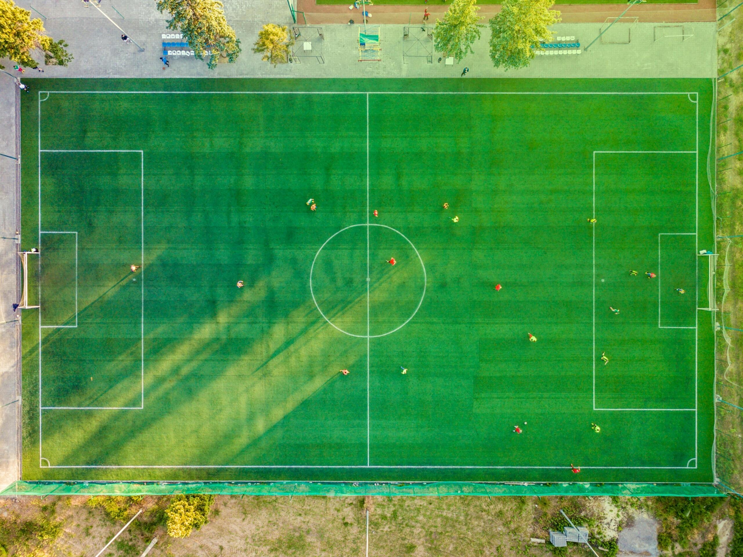 A bird’s-eye view of a soccer field showing clearly marked lines, goals at both ends, and players positioned across the pitch during a match.