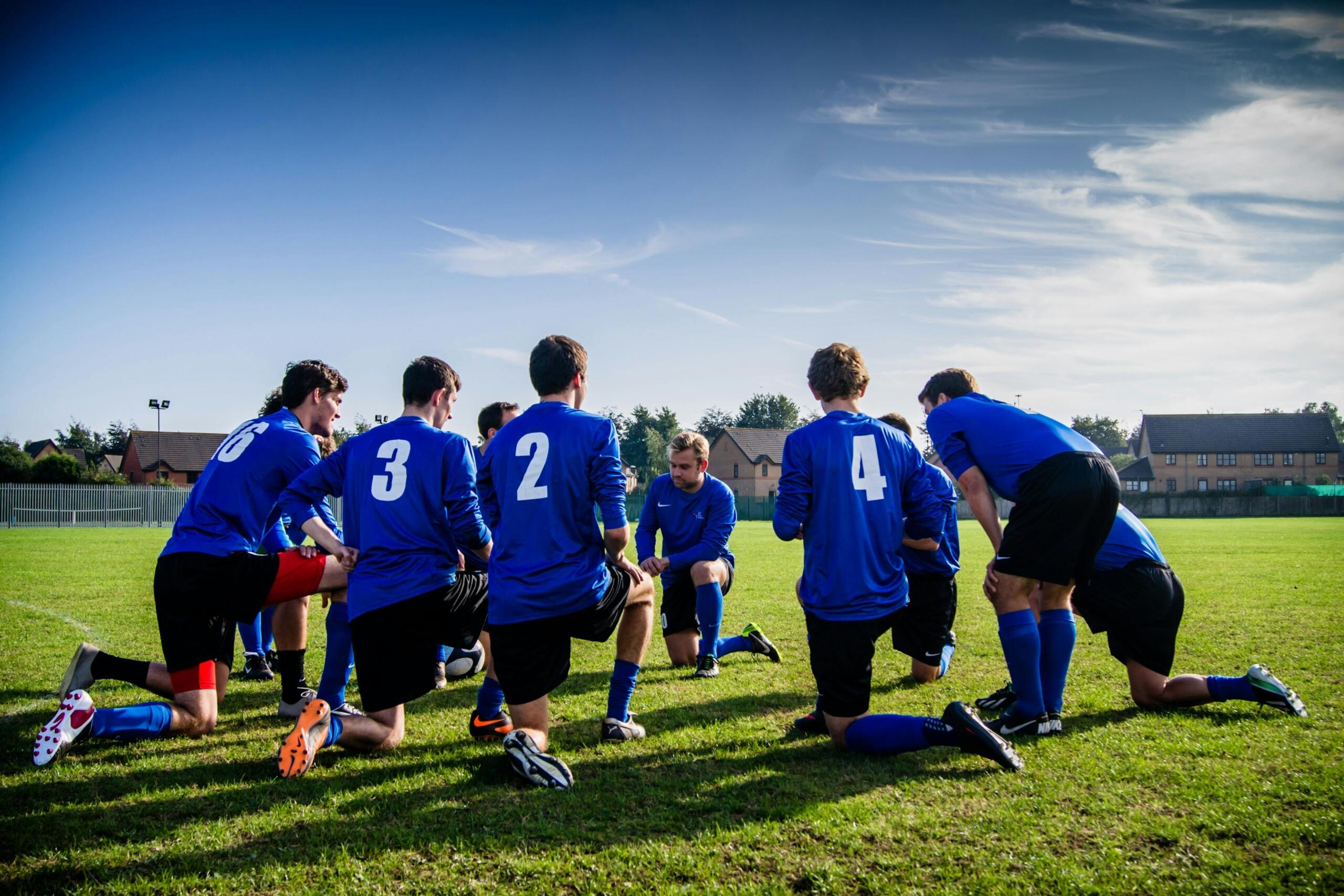 A group of soccer players in blue jerseys gathered around their coach on a grassy field, listening attentively as the coach gives instructions.