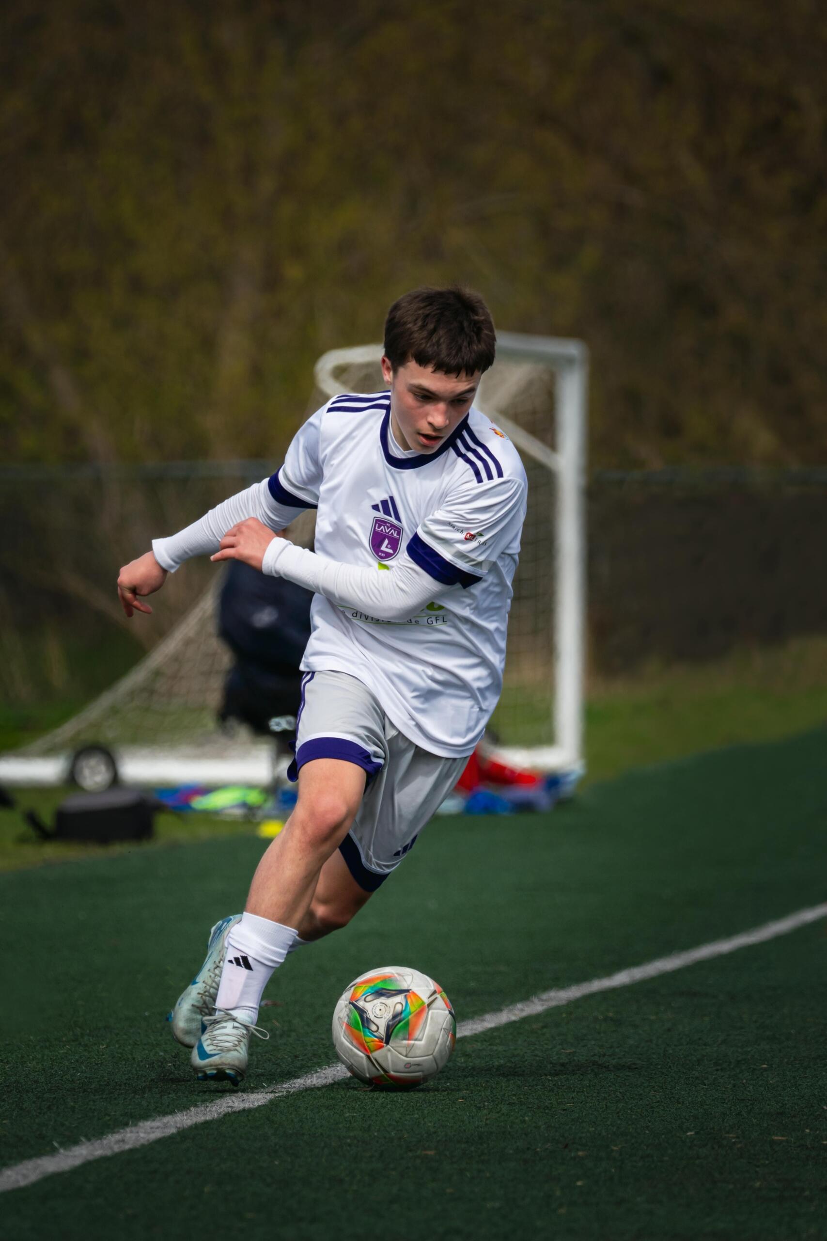 A teen in a white soccer uniform skillfully kicks the ball mid-action on a green field, demonstrating focus and athleticism.