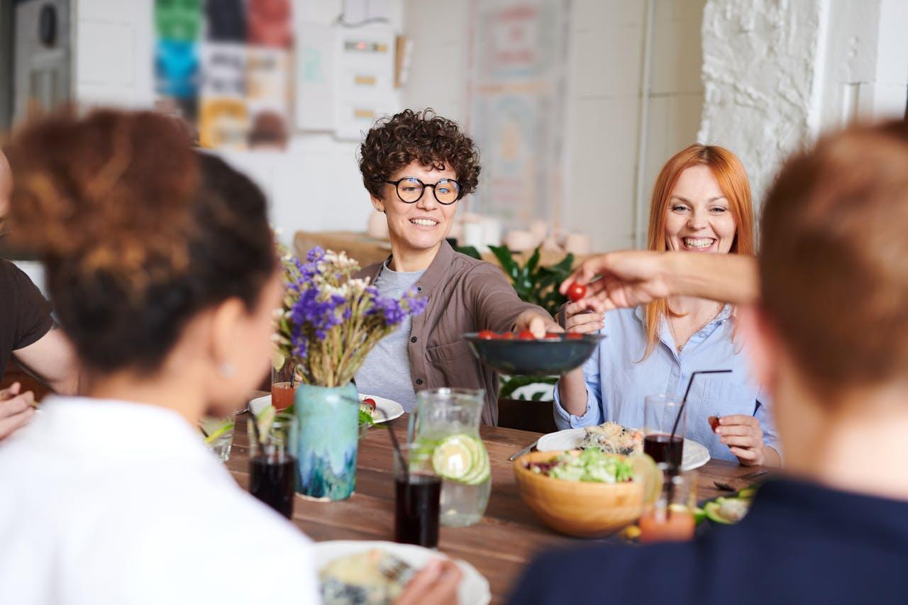A group of people enjoying a meal together at a table, with salads, drinks, and a vase of purple flowers in a cozy setting.