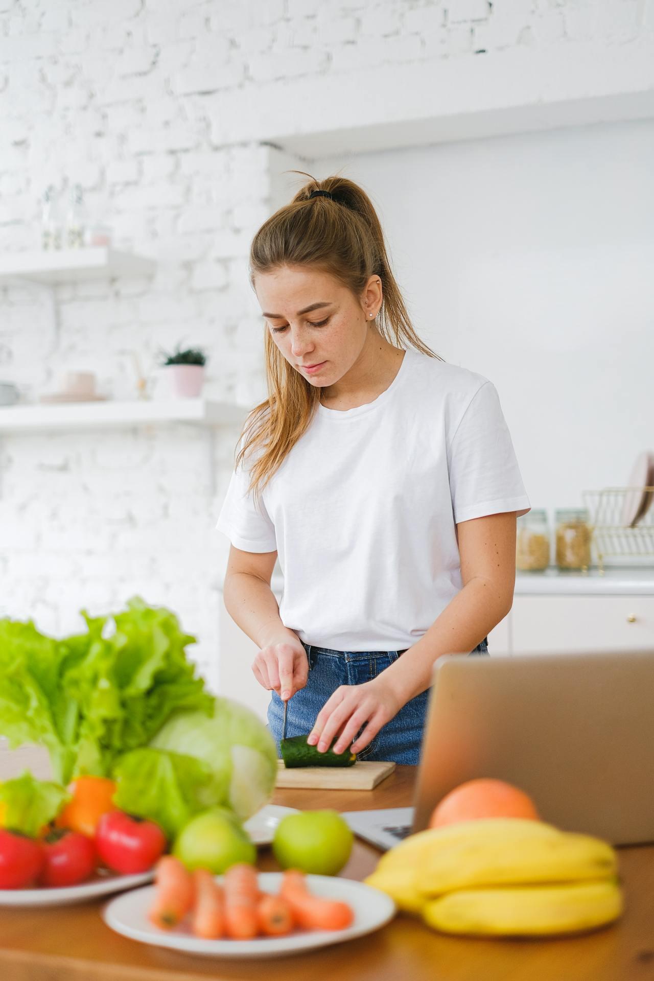A person in a white t-shirt chops a cucumber at a kitchen table filled with fresh vegetables and fruits. A laptop is nearby.