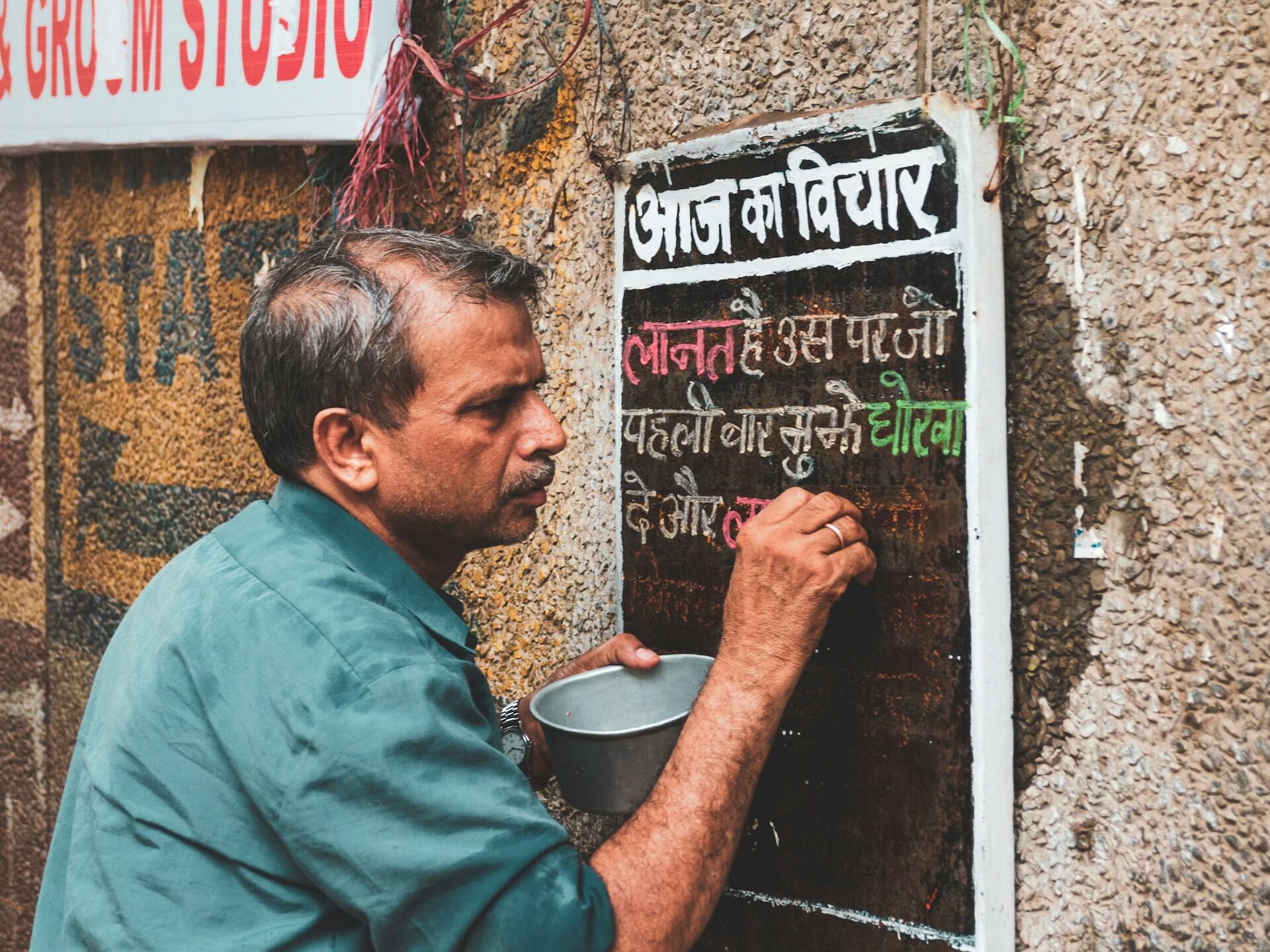 Man in green shirt writing colorful Hindi text on a blackboard mounted on a textured wall outdoors.