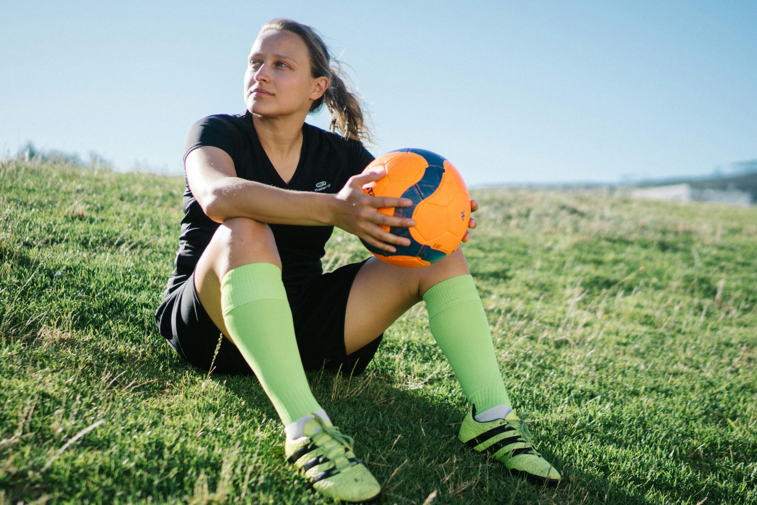 woman in a soccer uniform sitting on the grass, smiling and holding a soccer ball in her hands.