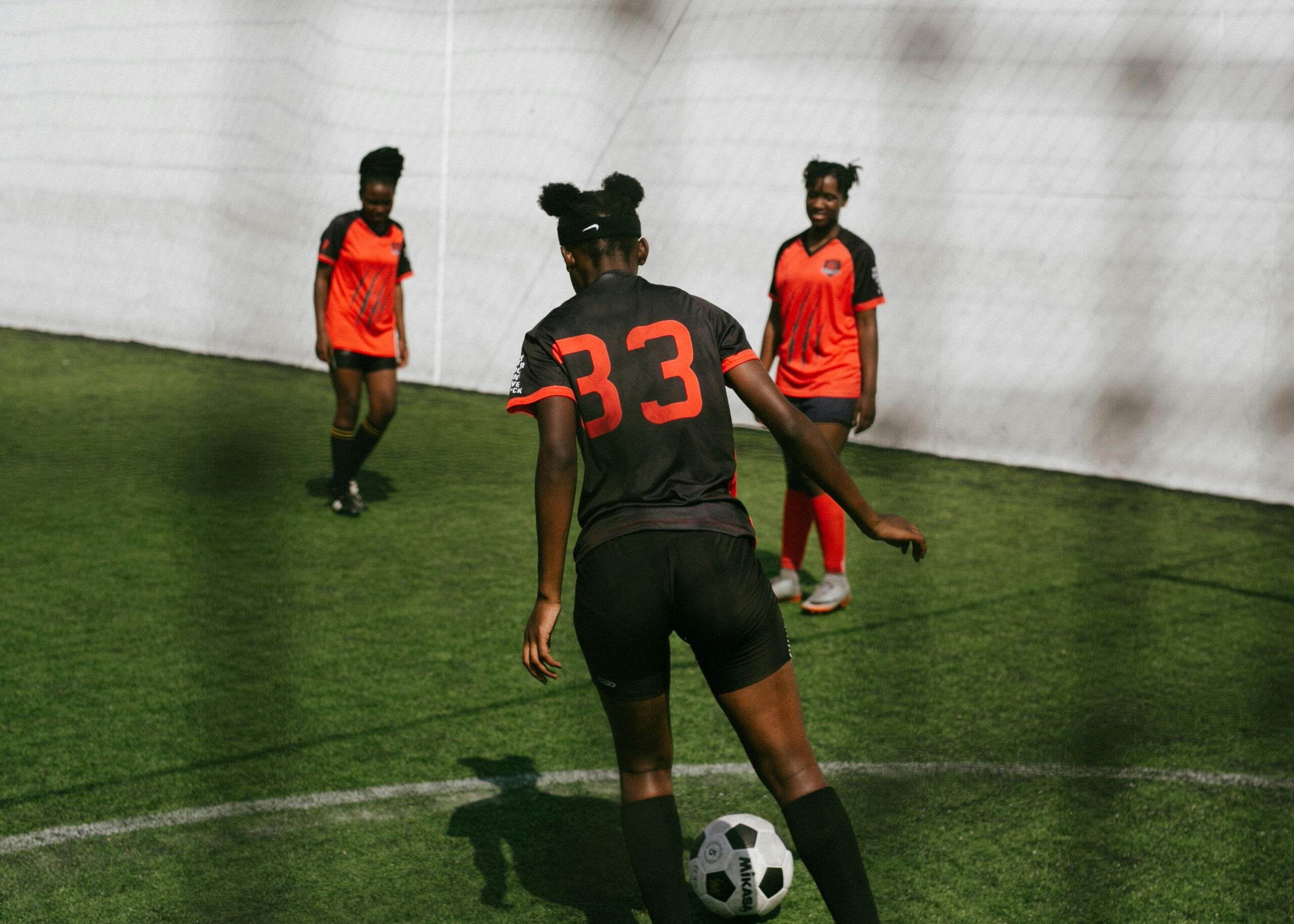 A view from behind the goal net showing three female soccer players on the field, captured mid-play as they move toward the ball with the net framing the scene.