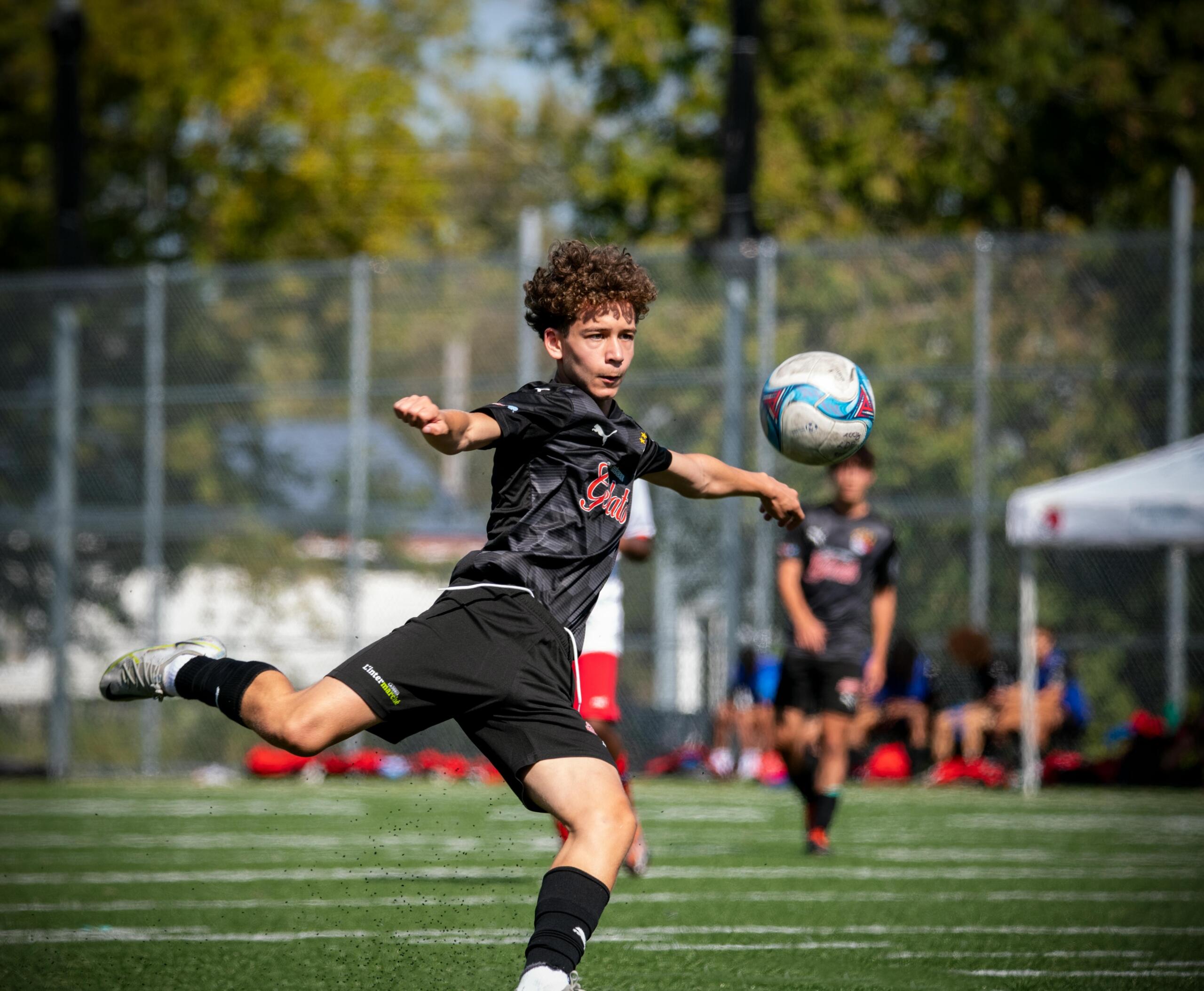 A male teenager in athletic clothing kicking a soccer ball on a grassy field, captured mid-action with focused energy and motion.
