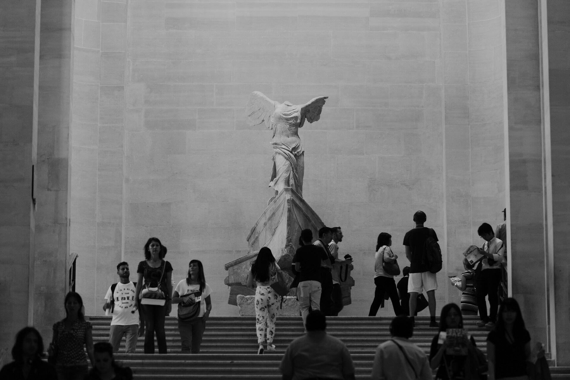 A group of visitors climbs stairs towards the iconic Winged Victory statue, surrounded by grand stone walls.