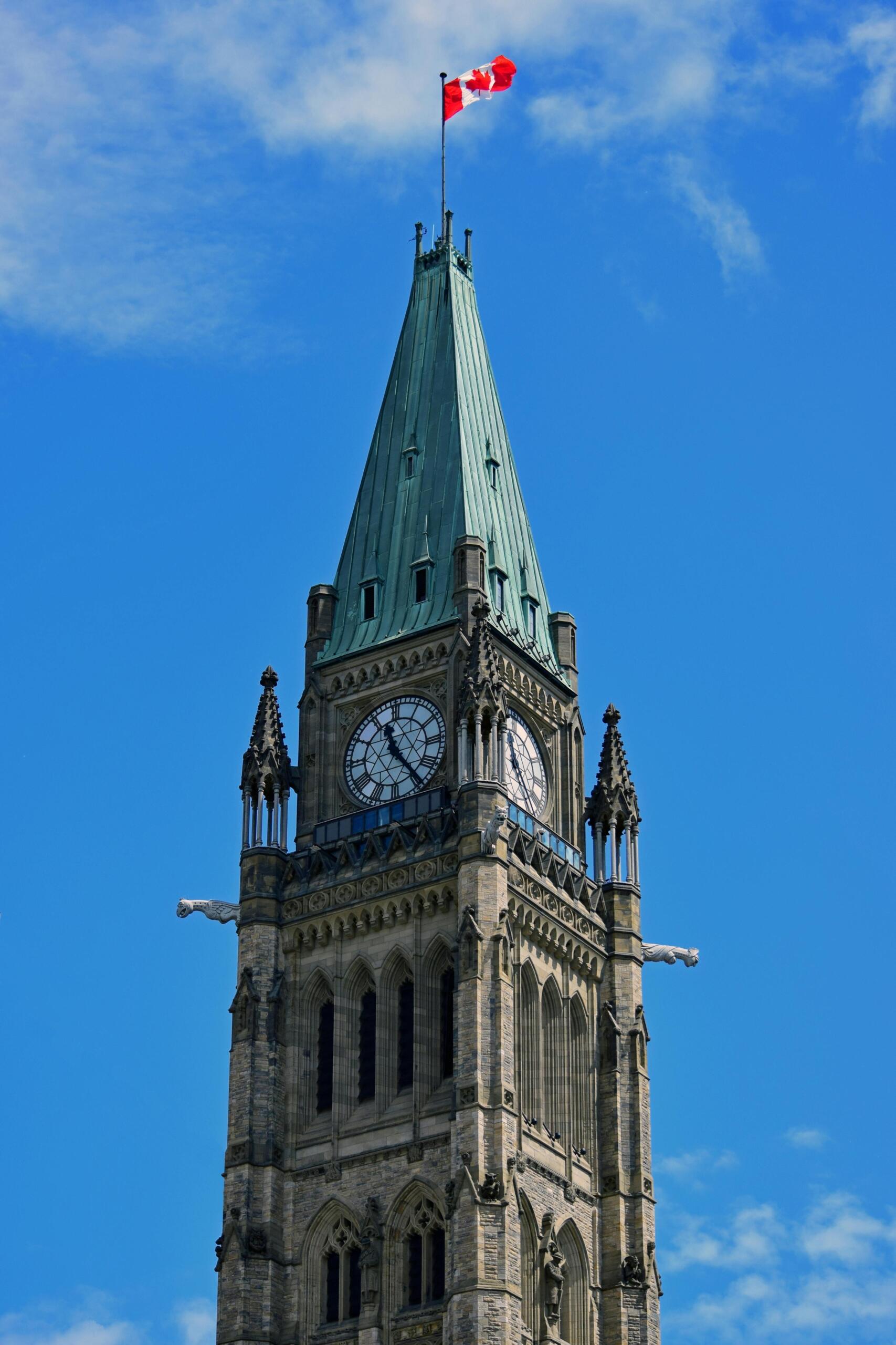 The iconic clock tower on Parliament Hill in Ottawa, standing tall against a clear sky with intricate stonework and the Canadian flag waving at the top.