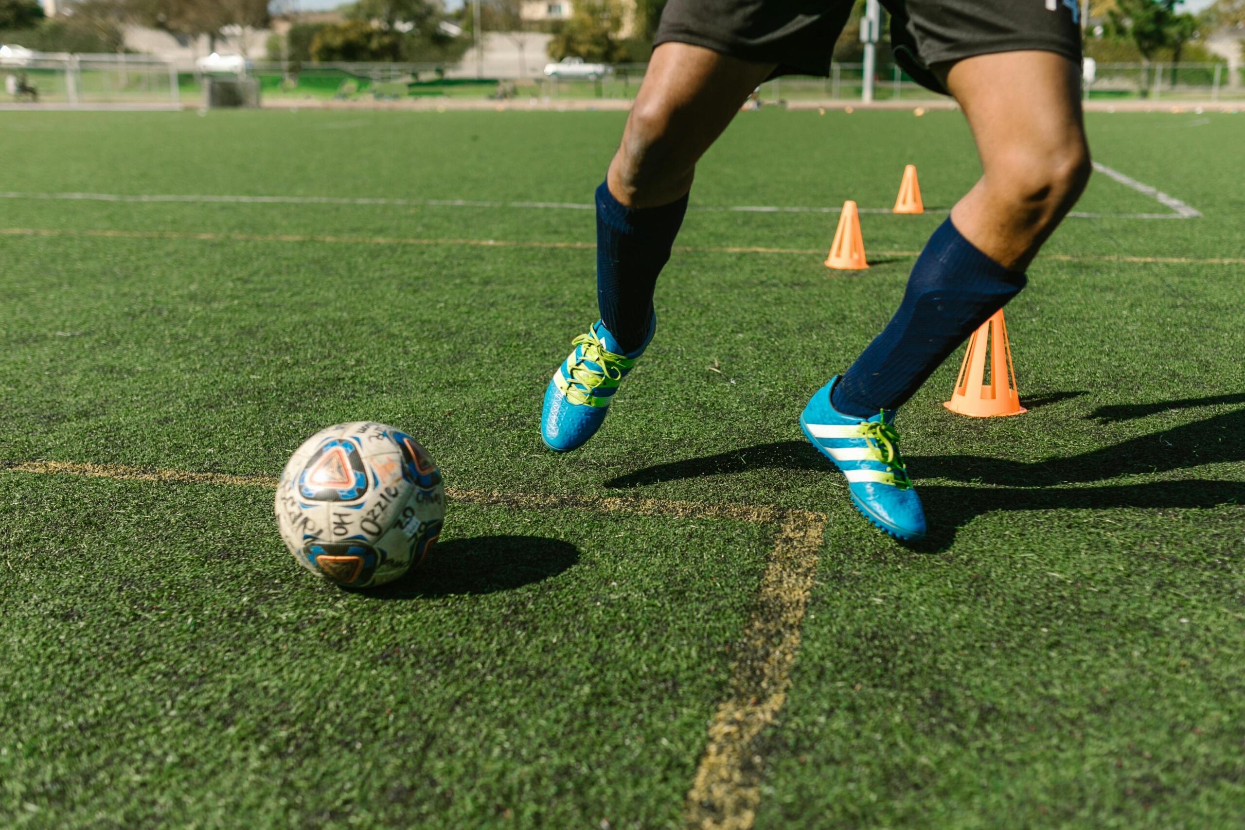 Close-up of a man's feet in cleats kicking a soccer ball on a grassy field, capturing the moment of contact between foot and ball.