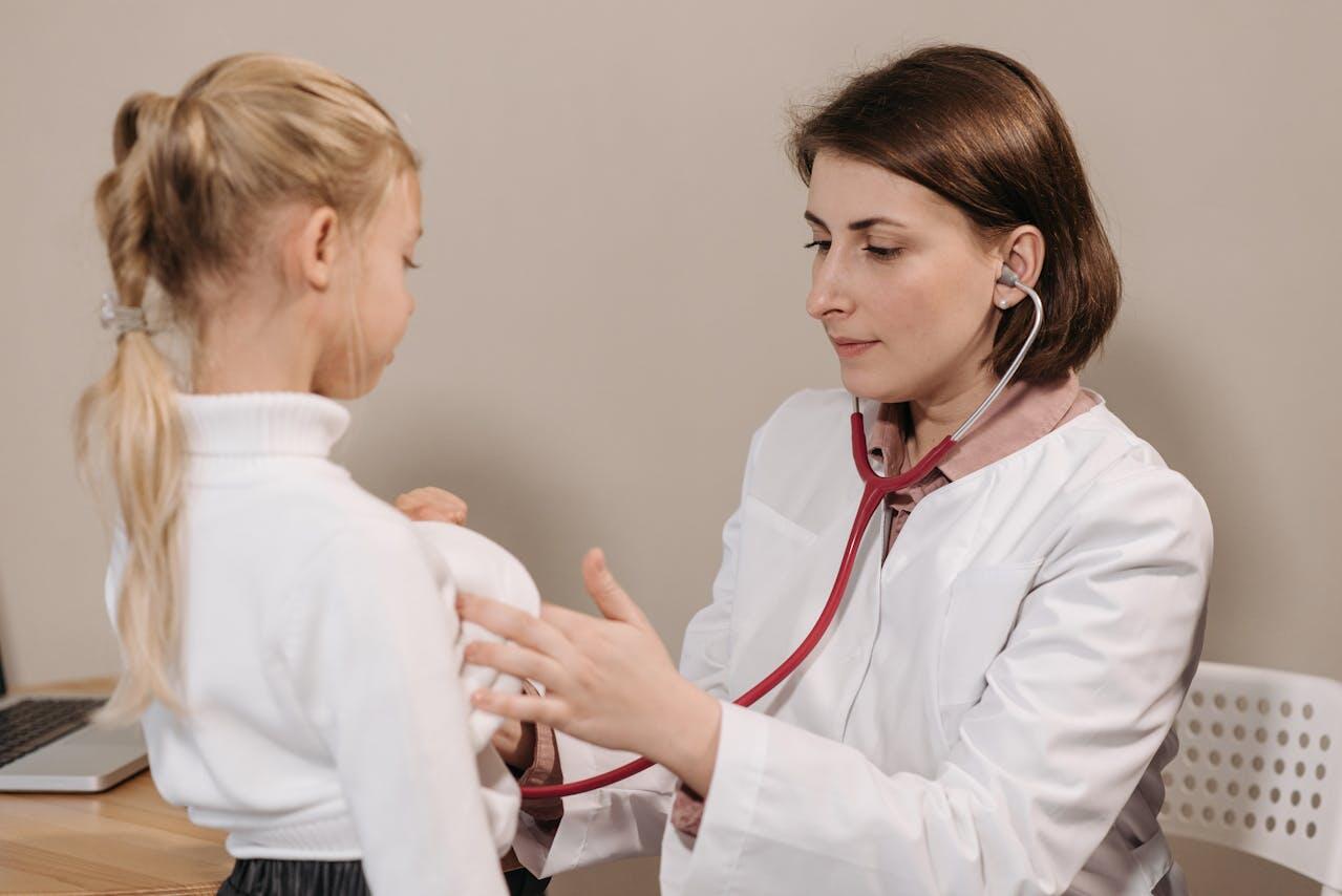 A doctor in a white coat examines a child, using a stethoscope and providing medical attention in a warmly lit office setting.
