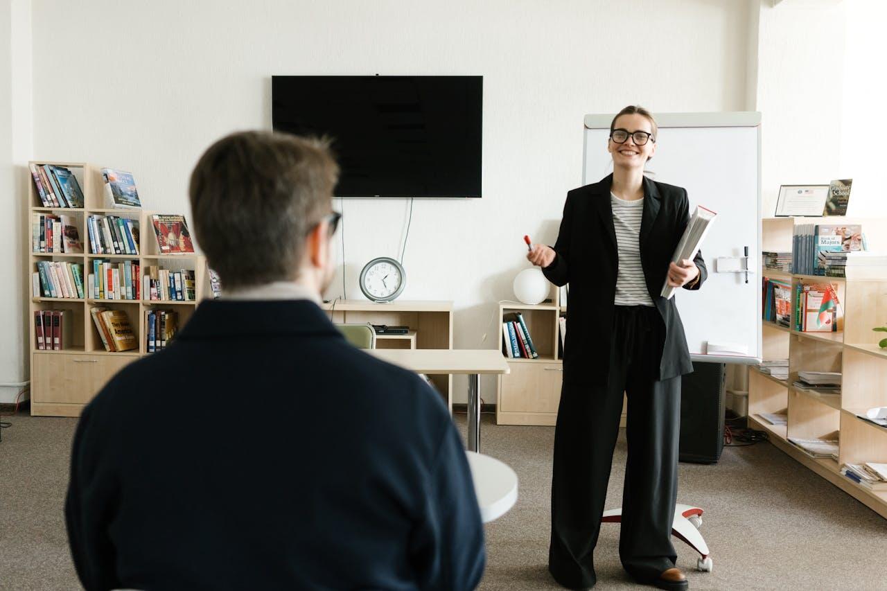 A person stands confidently in front of a classroom, presenting with notes, while a seated individual listens attentively. Bookshelves are visible.