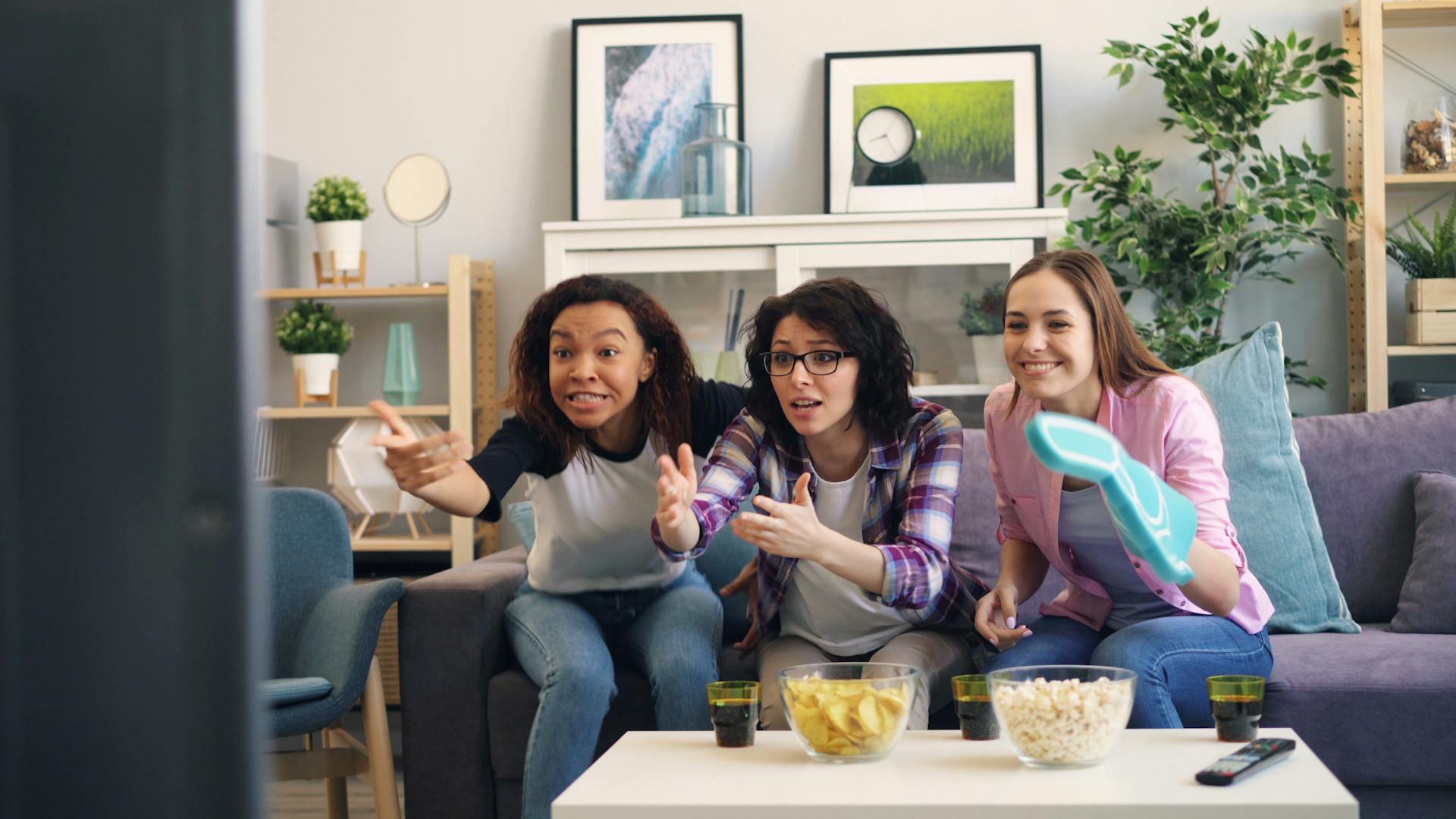 Three friends are excitedly watching TV together on a couch, surrounded by snacks and drinks in a cozy living room setting.