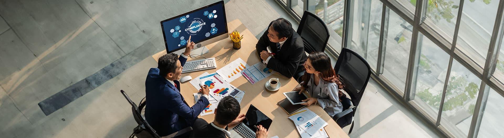Overhead view of a diverse business team in a modern office discussing compliance and sustainability metrics, with charts, digital devices, and a large window providing natural light.