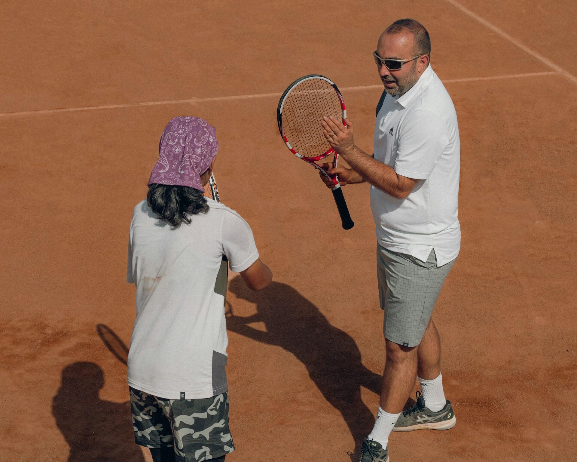 A tennis coach demonstrates a technique to a player on a clay court, both holding rackets and engaged in the lesson.