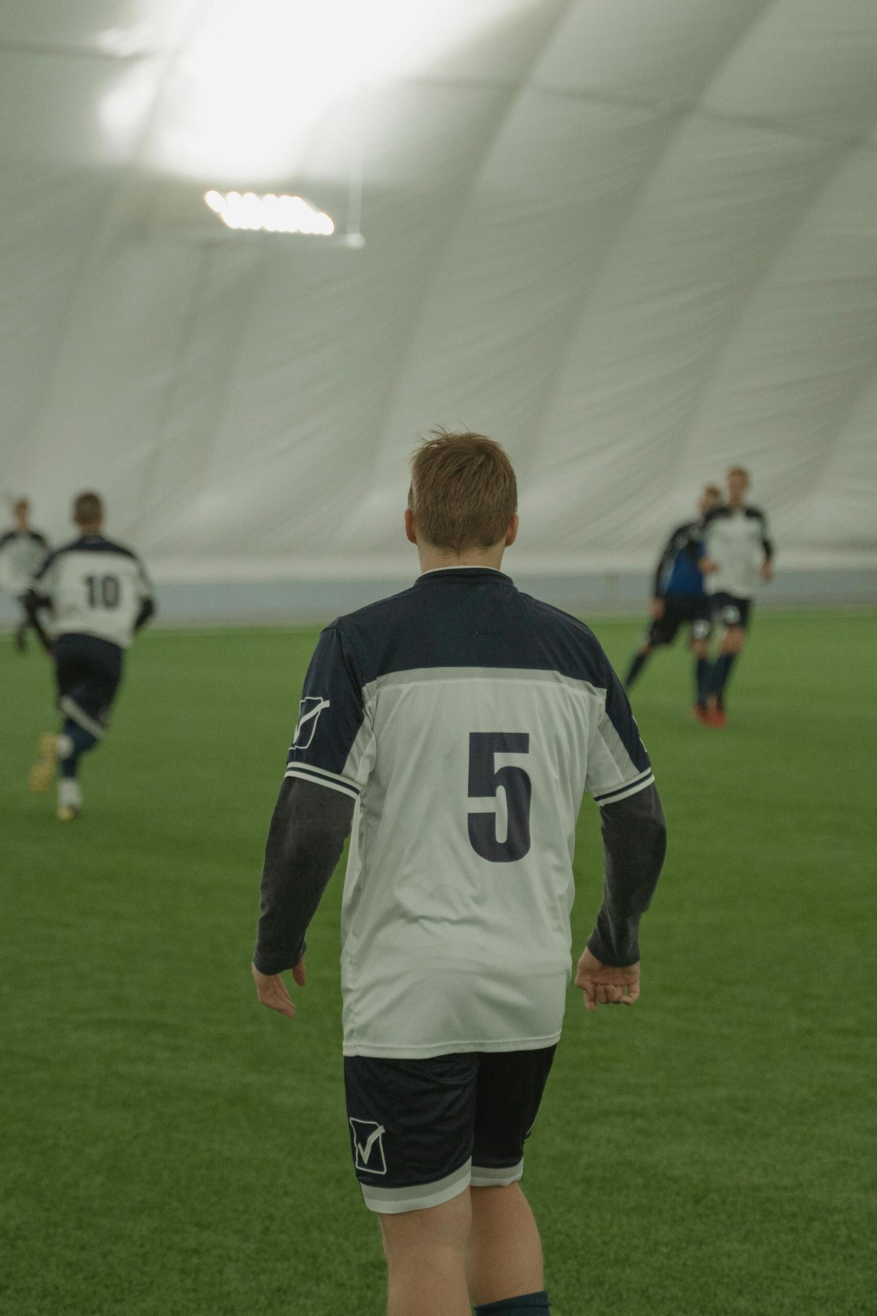 A group of soccer players actively playing on an indoor soccer field, surrounded by netted walls and artificial turf under overhead lights.