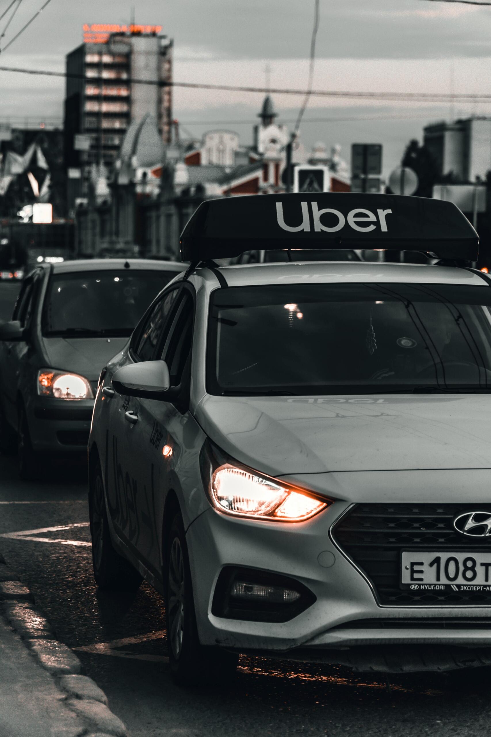 A silver Uber car with headlights on waits in evening traffic on a city street, symbolizing gig economy transportation services.