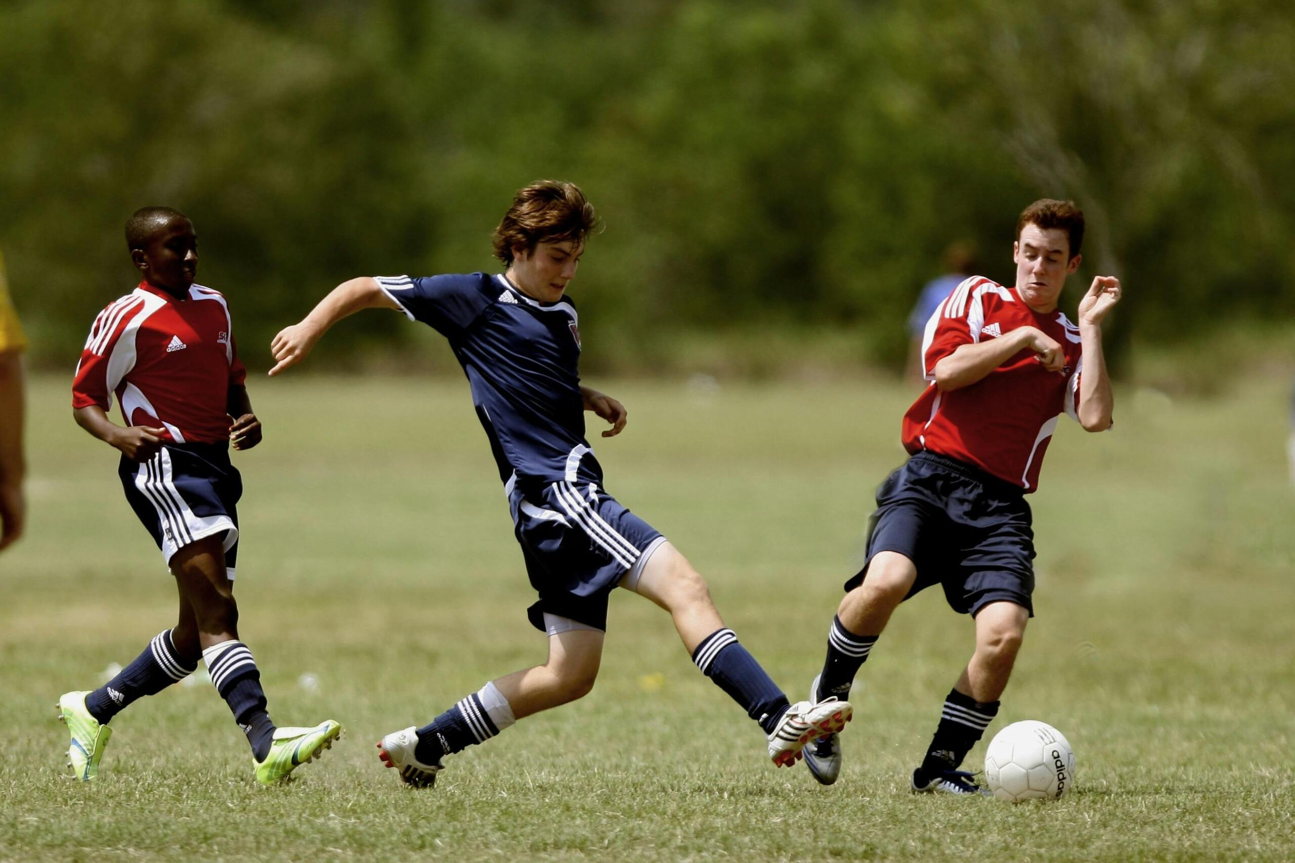 A group of teens energetically playing soccer on a grassy field, actively chasing the ball and engaging in a fmatch in a field surrounded by forests.