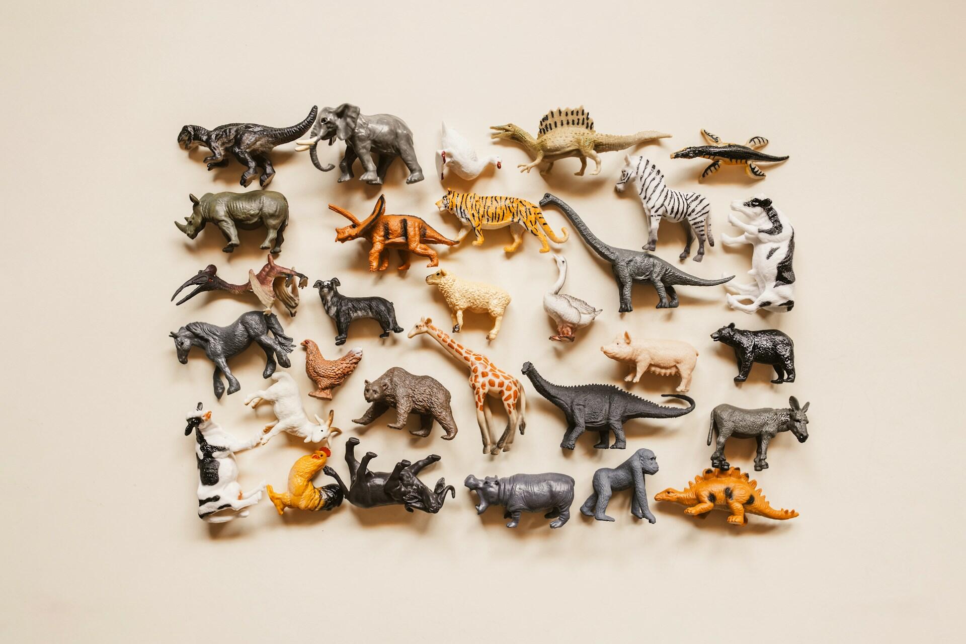 A colorful arrangement of various plastic animal figurines, including dinosaurs, mammals, and birds, displayed on a light background.