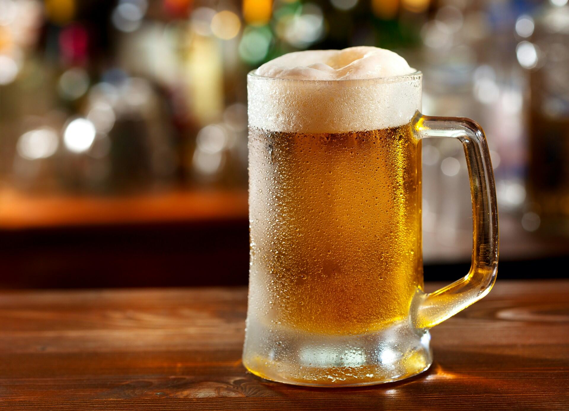 A chilled glass mug of golden beer with a frothy white head, sitting on a wooden bar against a blurred background.