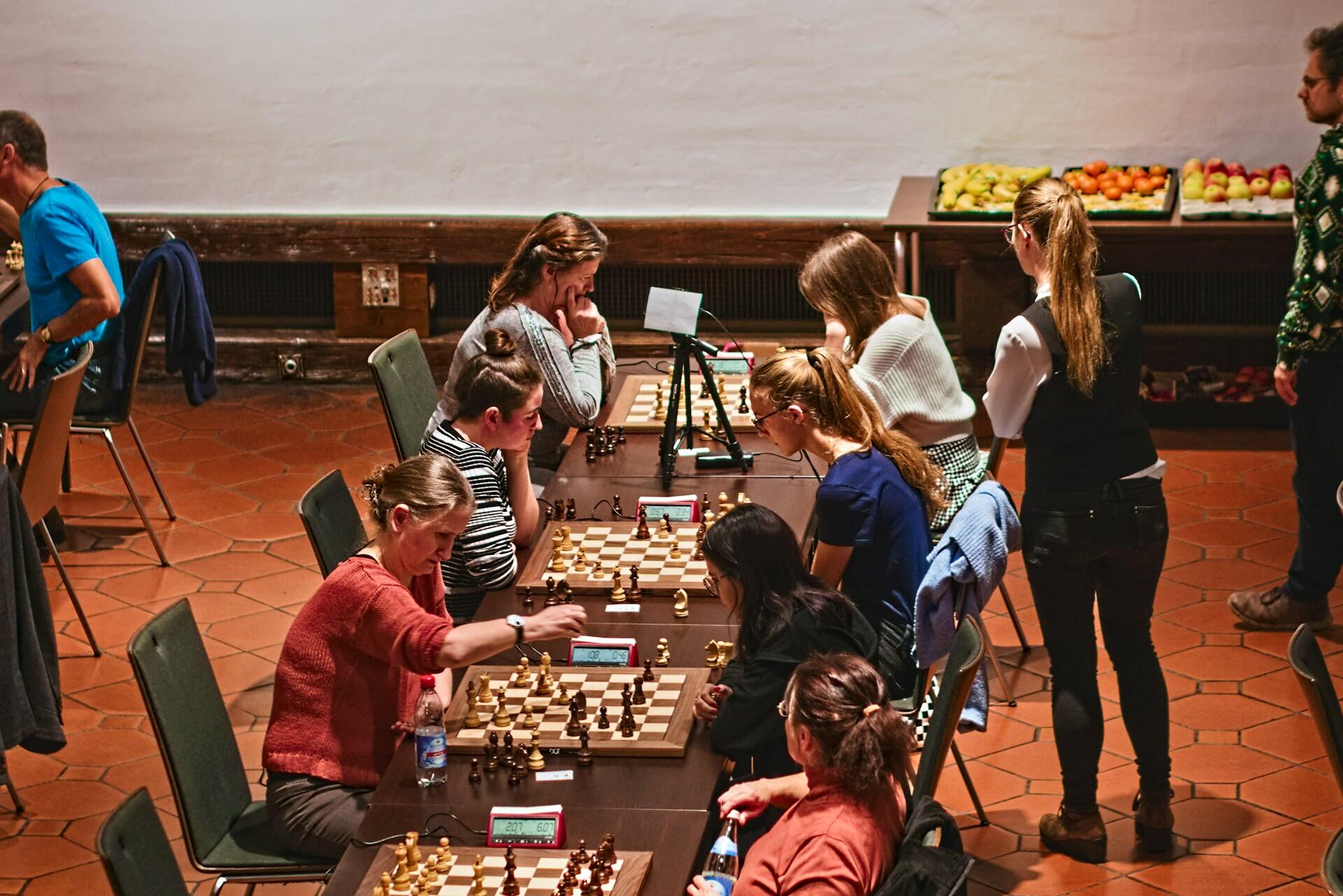 Eight individuals engage in a chess tournament at a long table, with clocks and chess pieces in a well-lit indoor setting.