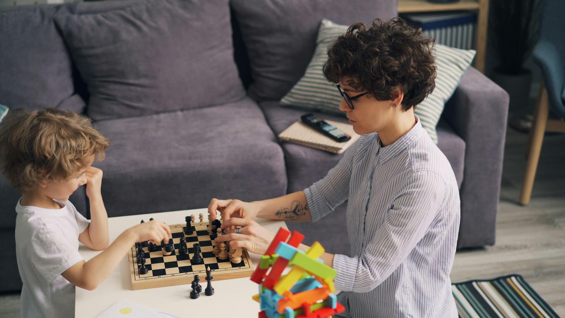 A child and an adult engage in a chess game at a table, with colorful building blocks nearby and a cozy sofa in the background.