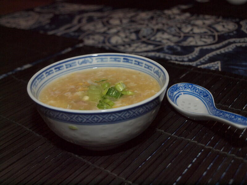 A bowl of soup with chopped green onions sits on a bamboo mat, accompanied by a small blue and white spoon.