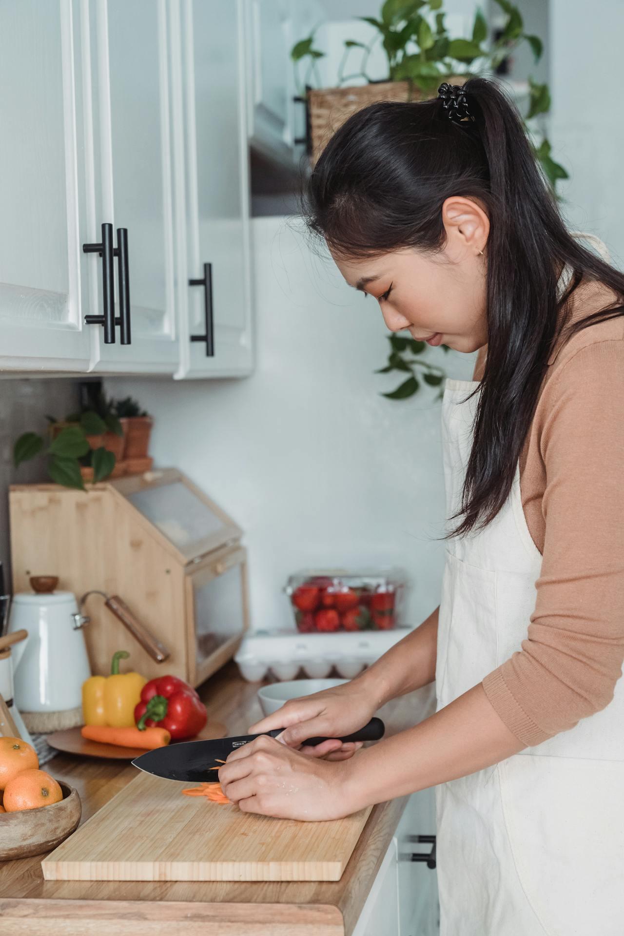 A person in an apron slices vegetables on a wooden cutting board in a bright kitchen filled with plants and fresh produce.
