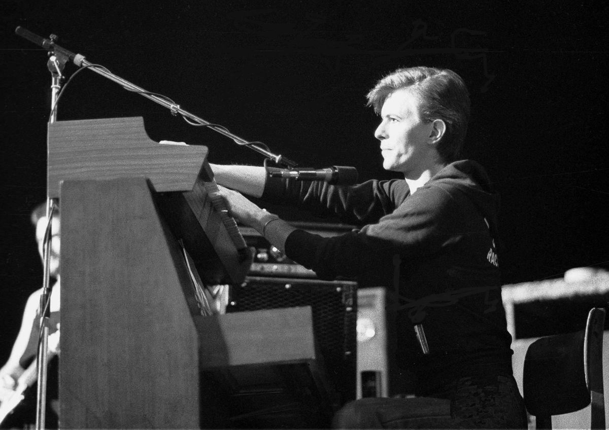 David Bowie sits by his piano with a microphone.